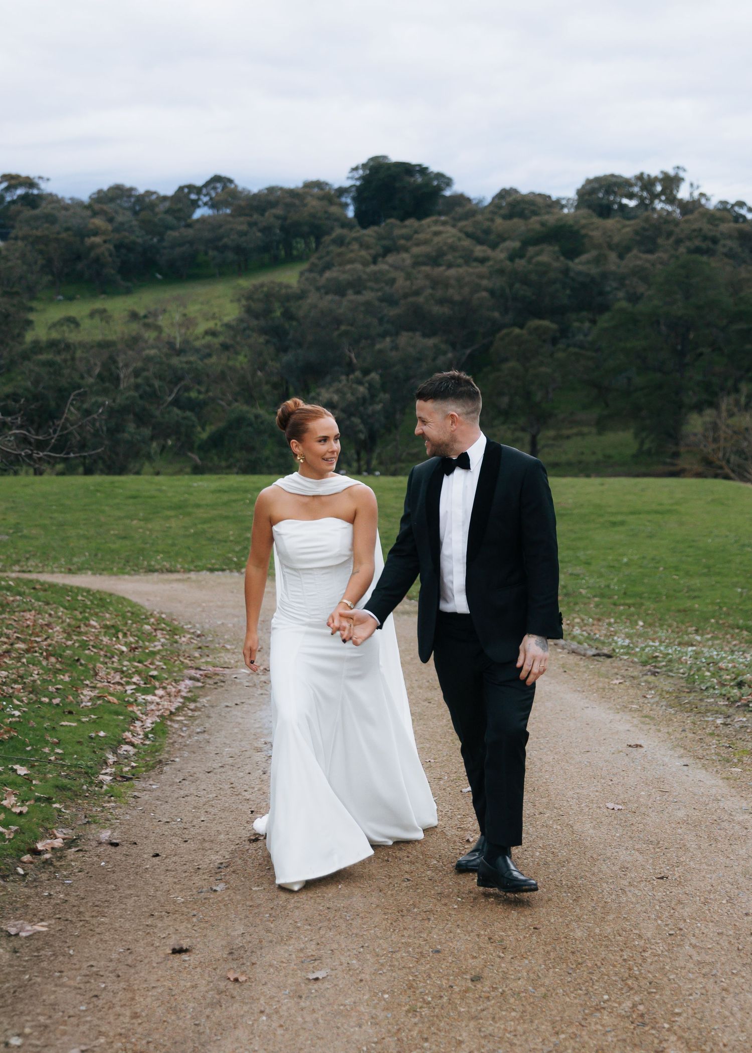 Bride in elegant white halter gown and groom in black tuxedo walking hand in hand on gravel path.