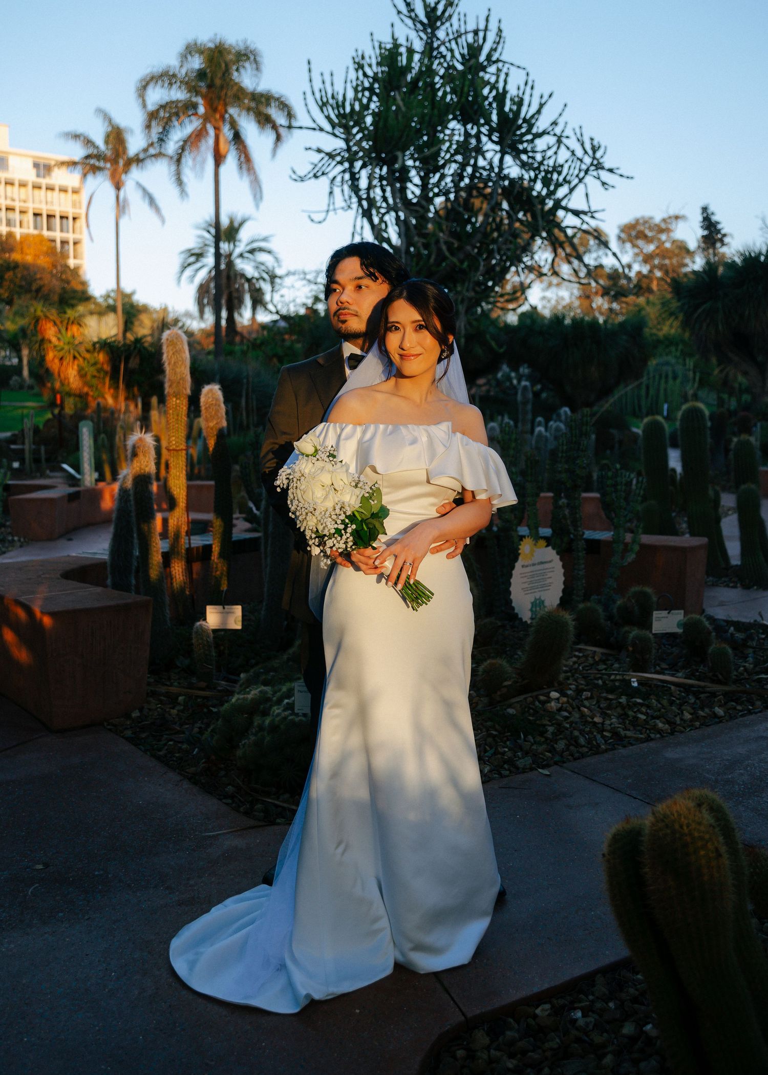 Bride and groom posing together in a desert garden with cacti and palm trees in warm golden hour light.