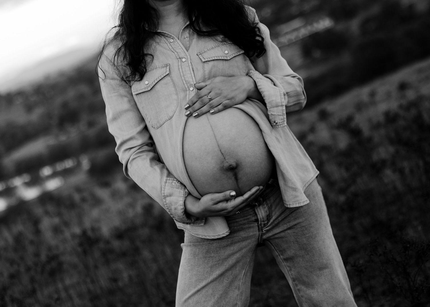 Black and white close-up of pregnant woman cradling bare baby bump in open field.