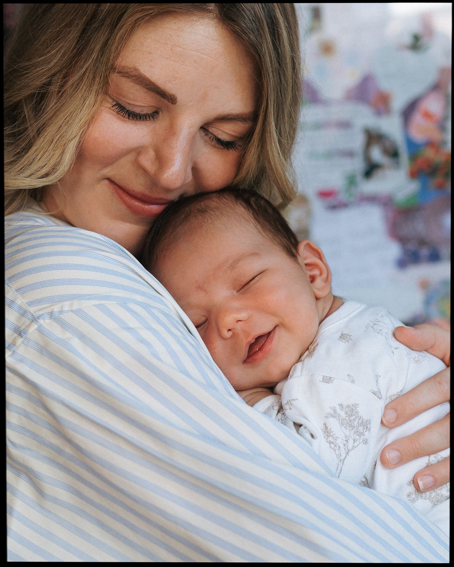Smiling mother in a blue striped shirt cradling a sleeping newborn baby in white against her chest in a warm, cozy home.