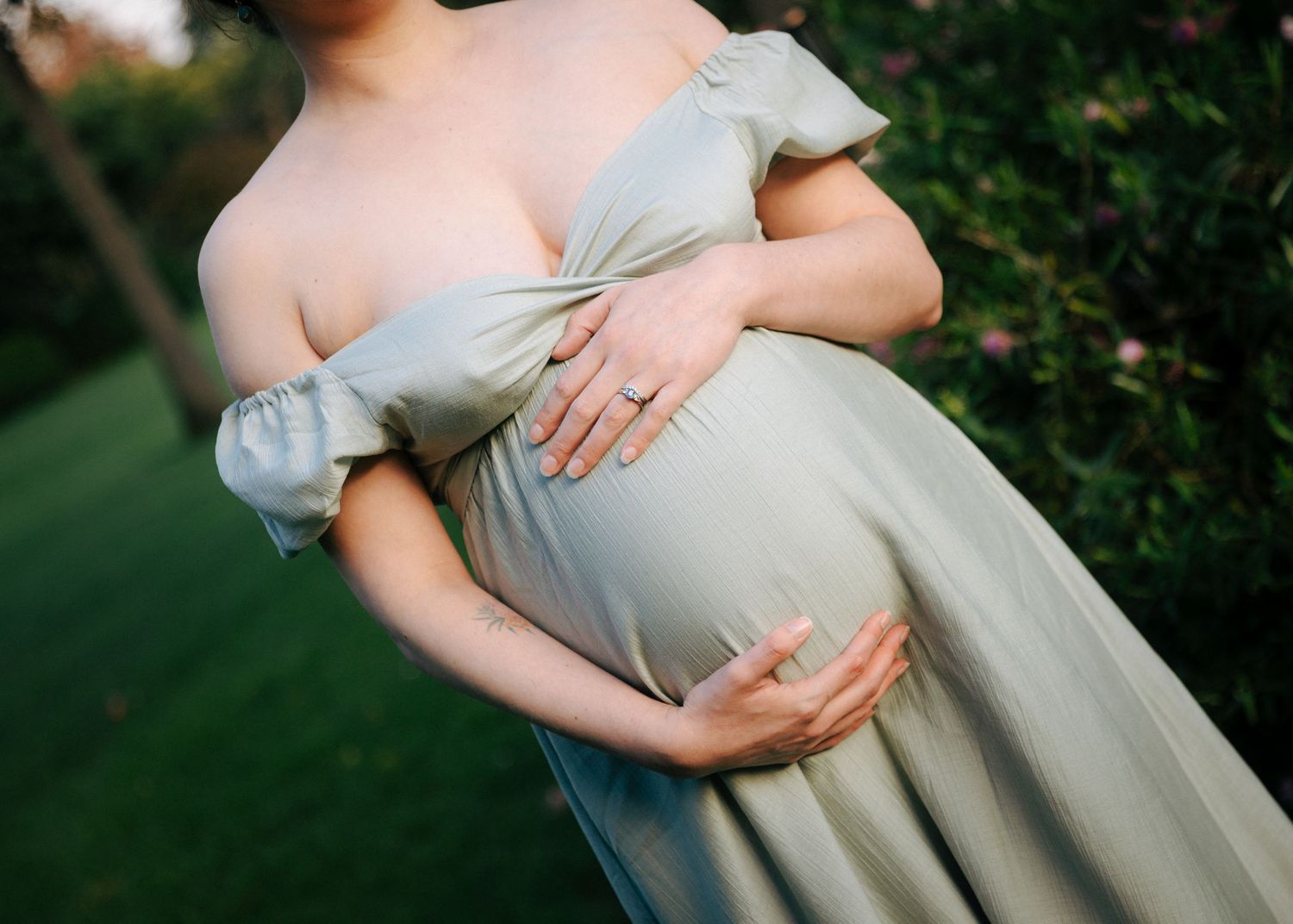 Close-up of pregnant belly in sage green off-shoulder maternity dress with partner's hands gently embracing.