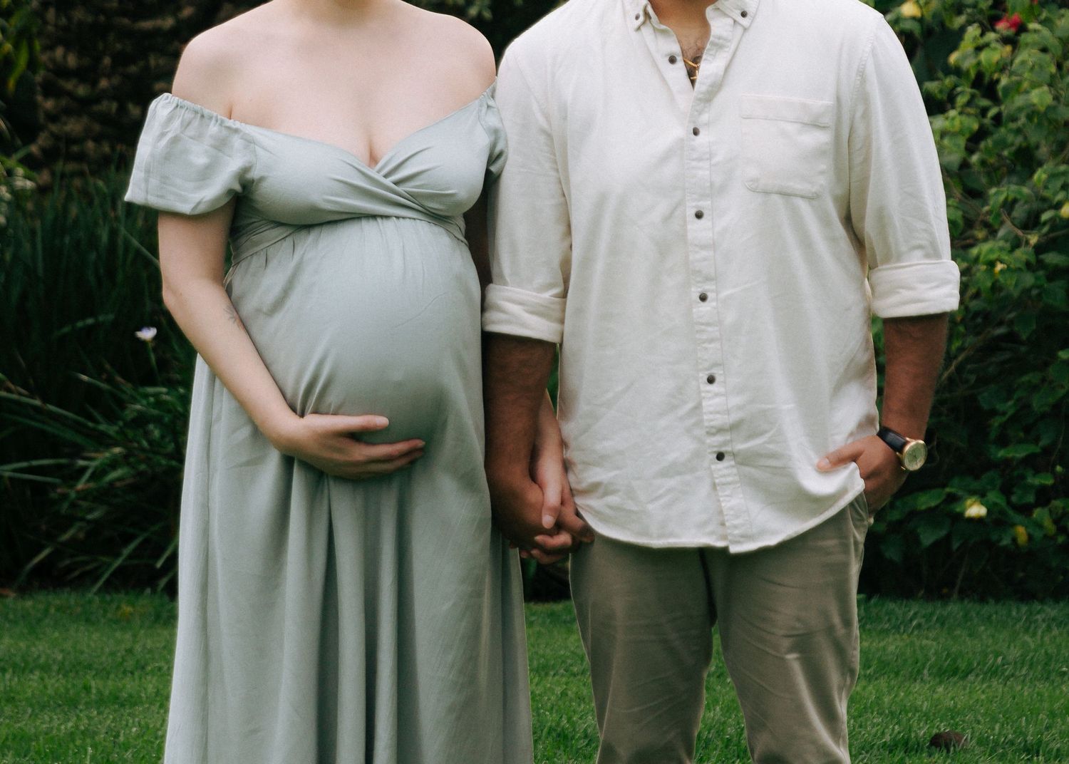 Pregnant couple holding hands in garden, woman in sage off-shoulder maternity dress showing baby bump.