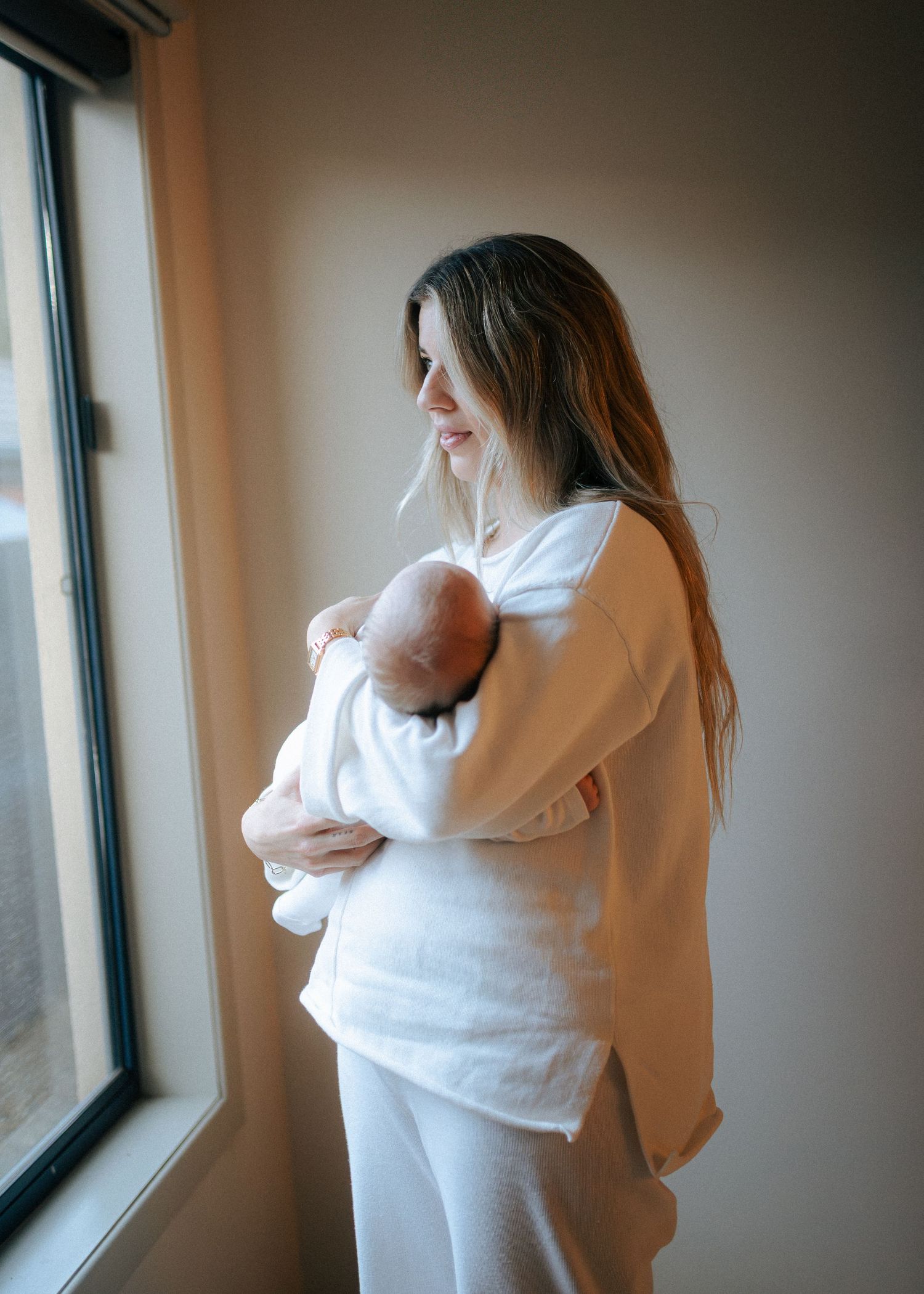 Mother in white linen outfit holding newborn baby while gazing out a window.