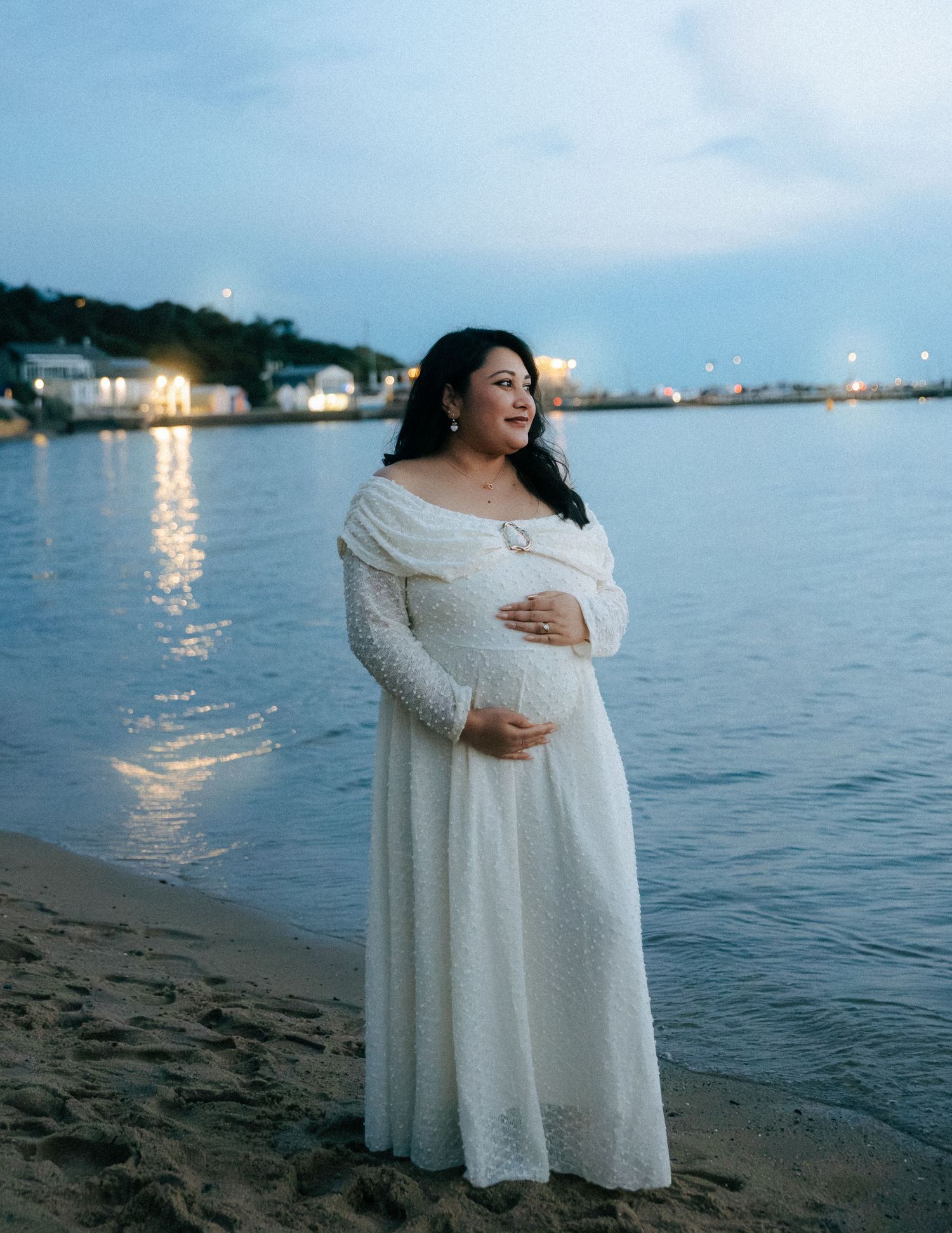 Pregnant woman in white lace off-shoulder gown posing on beach at twilight with harbor lights.
