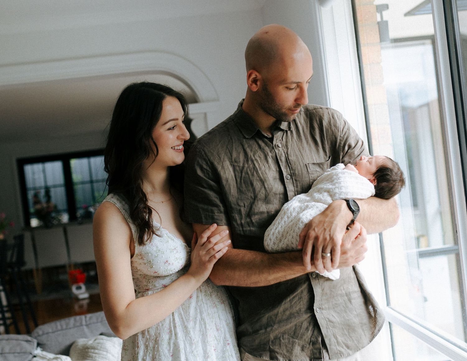 Couple standing by window smiling at newborn baby cradled in fathers arms at home.