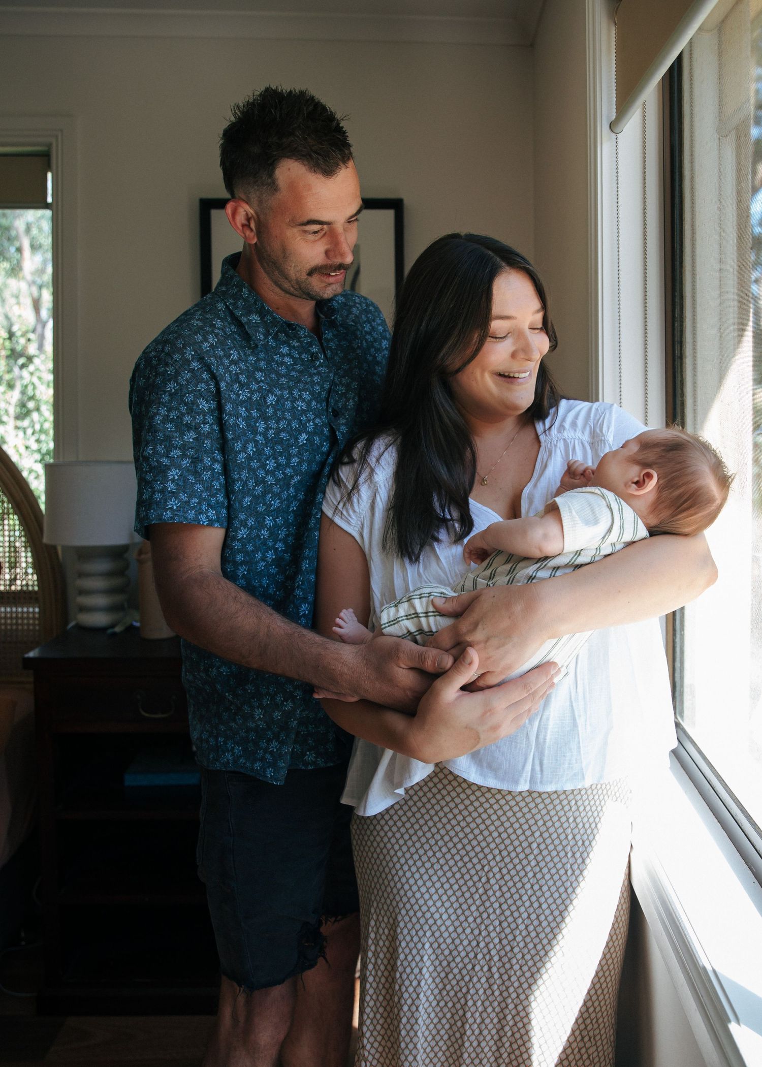 Happy parents standing by a window holding their newborn baby at home.