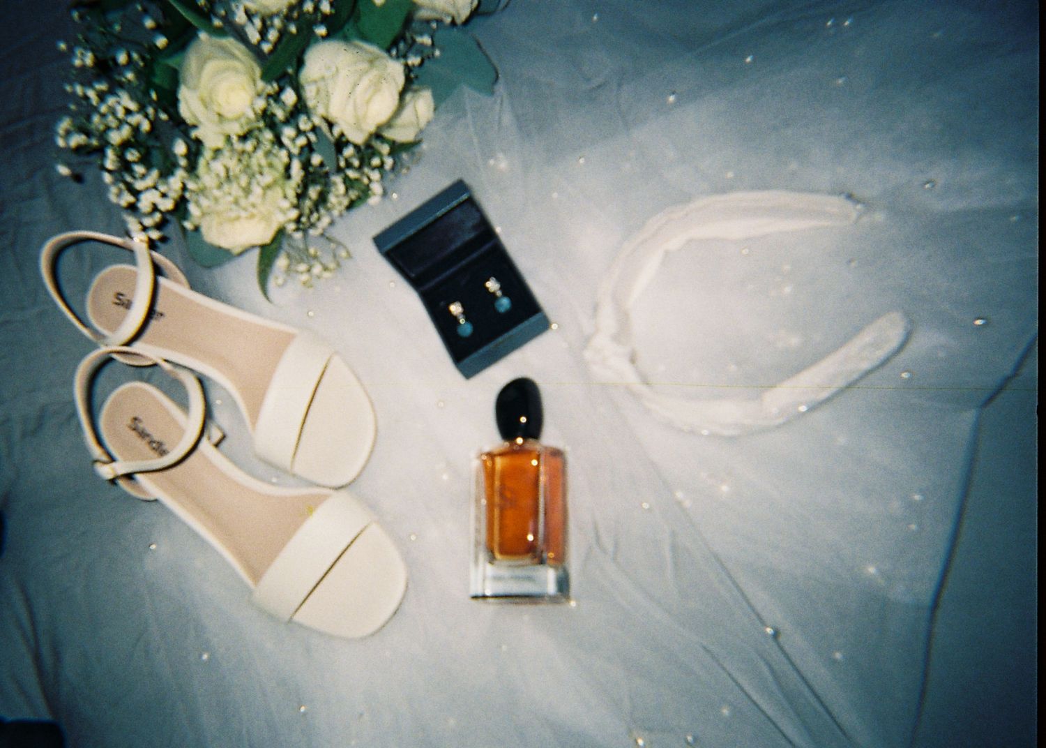 Bridal flat lay featuring white sandals, perfume, earrings, floral bouquet, and veil on tulle fabric.