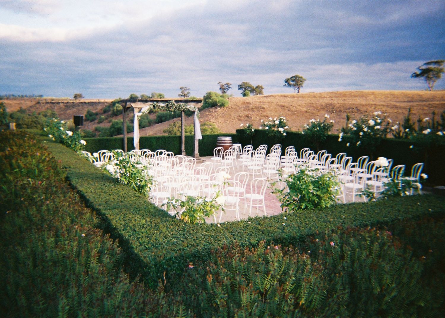 Outdoor wedding ceremony setup with white chairs arranged in rows surrounded by lush hedges.