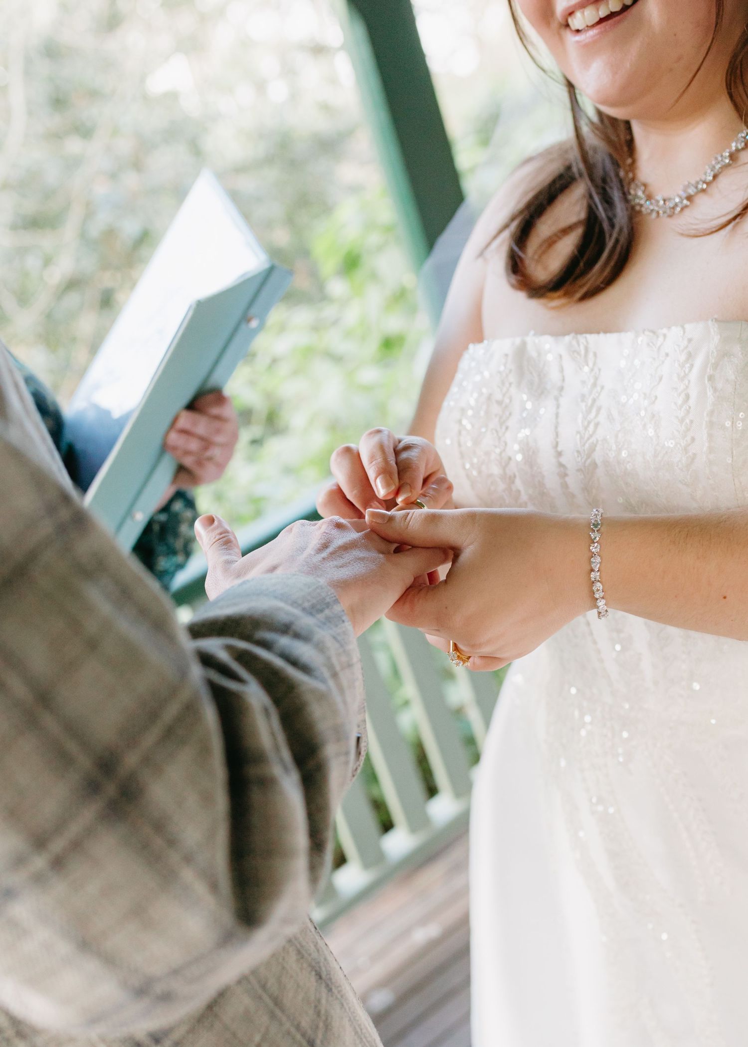 Bride smiling as she places a wedding ring on her groom's finger during an outdoor ceremony.