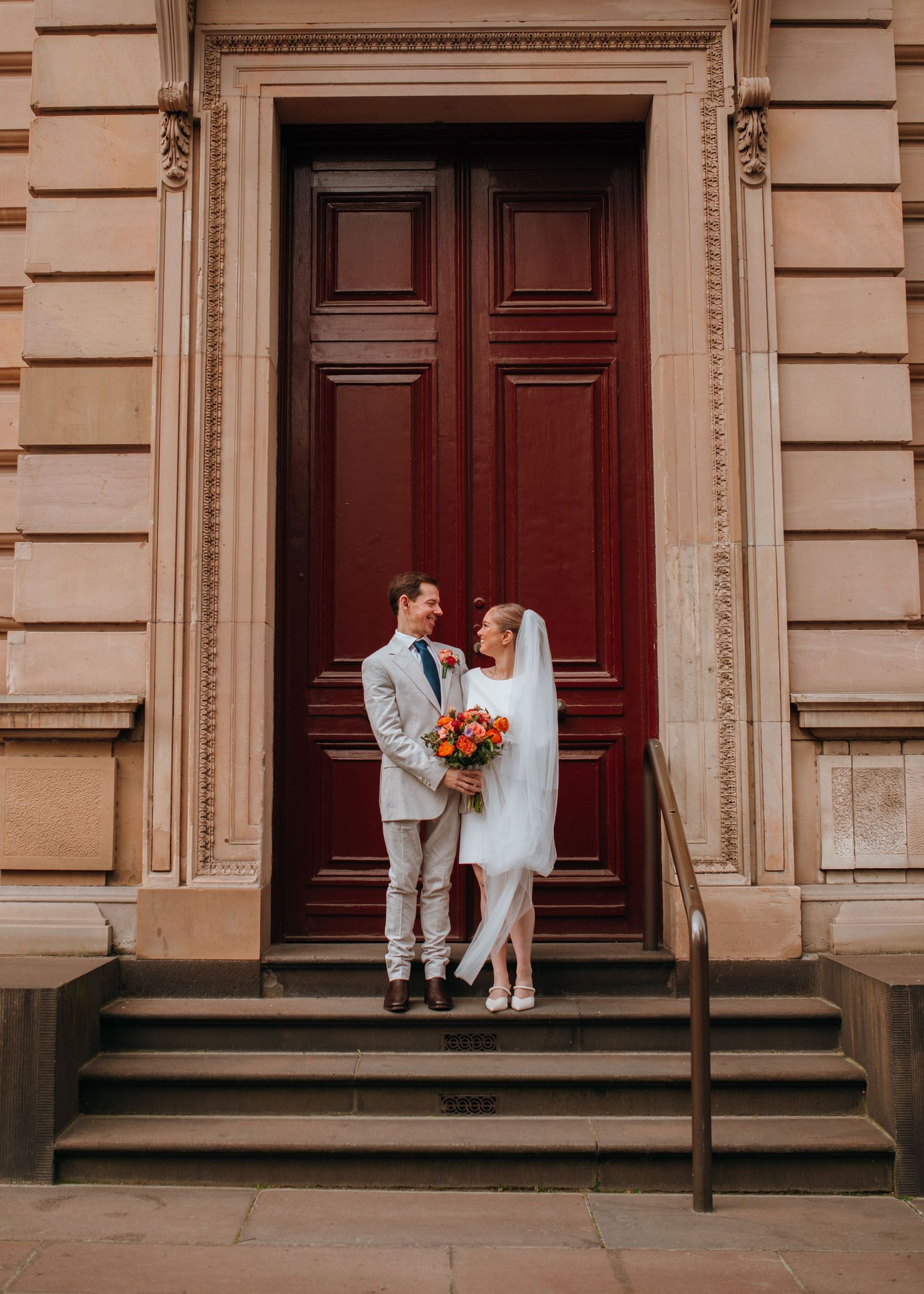 Bride in short white dress and groom in tan suit pose before grand red doors.