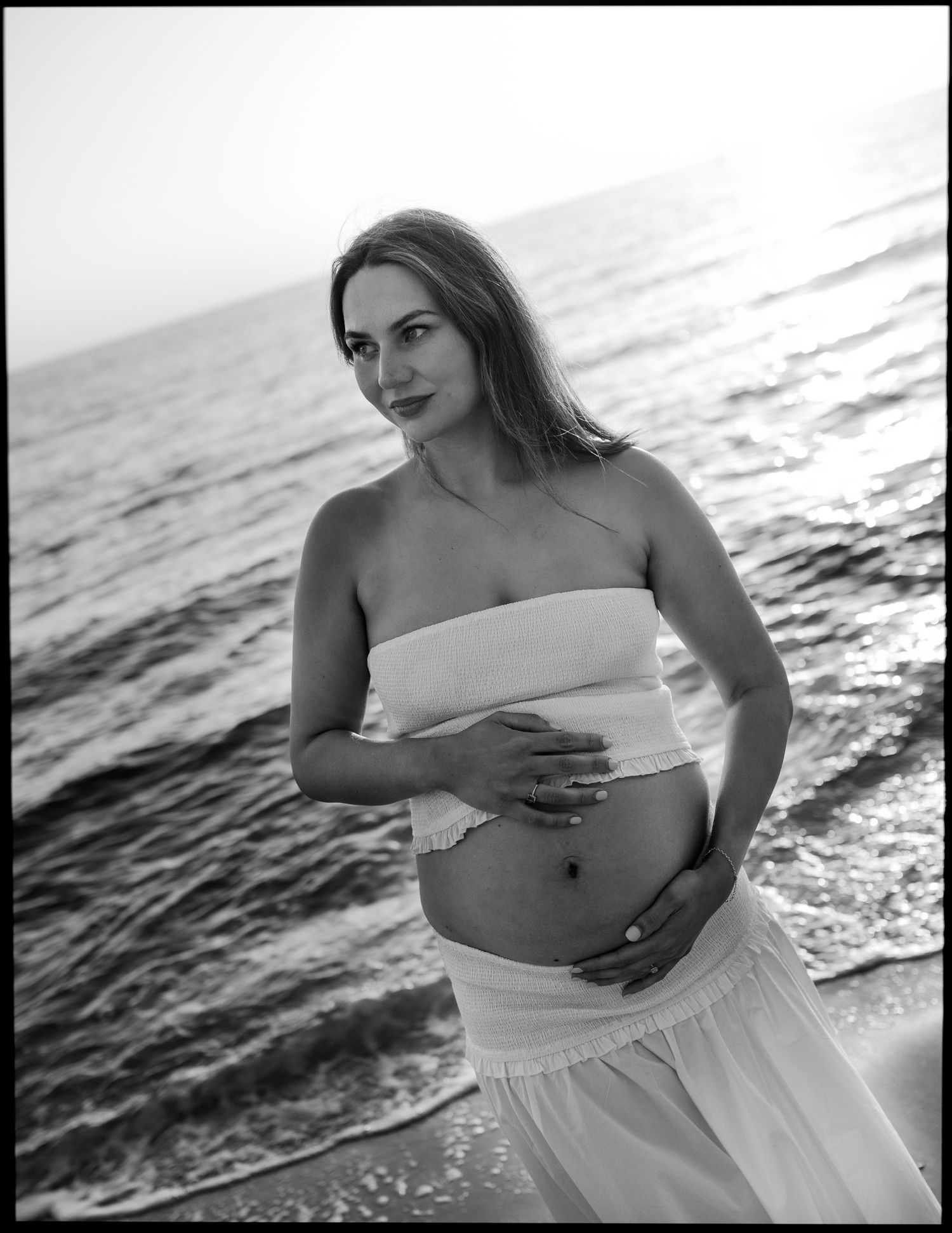 Pregnant woman in white tube top cradling baby bump on ocean beach.