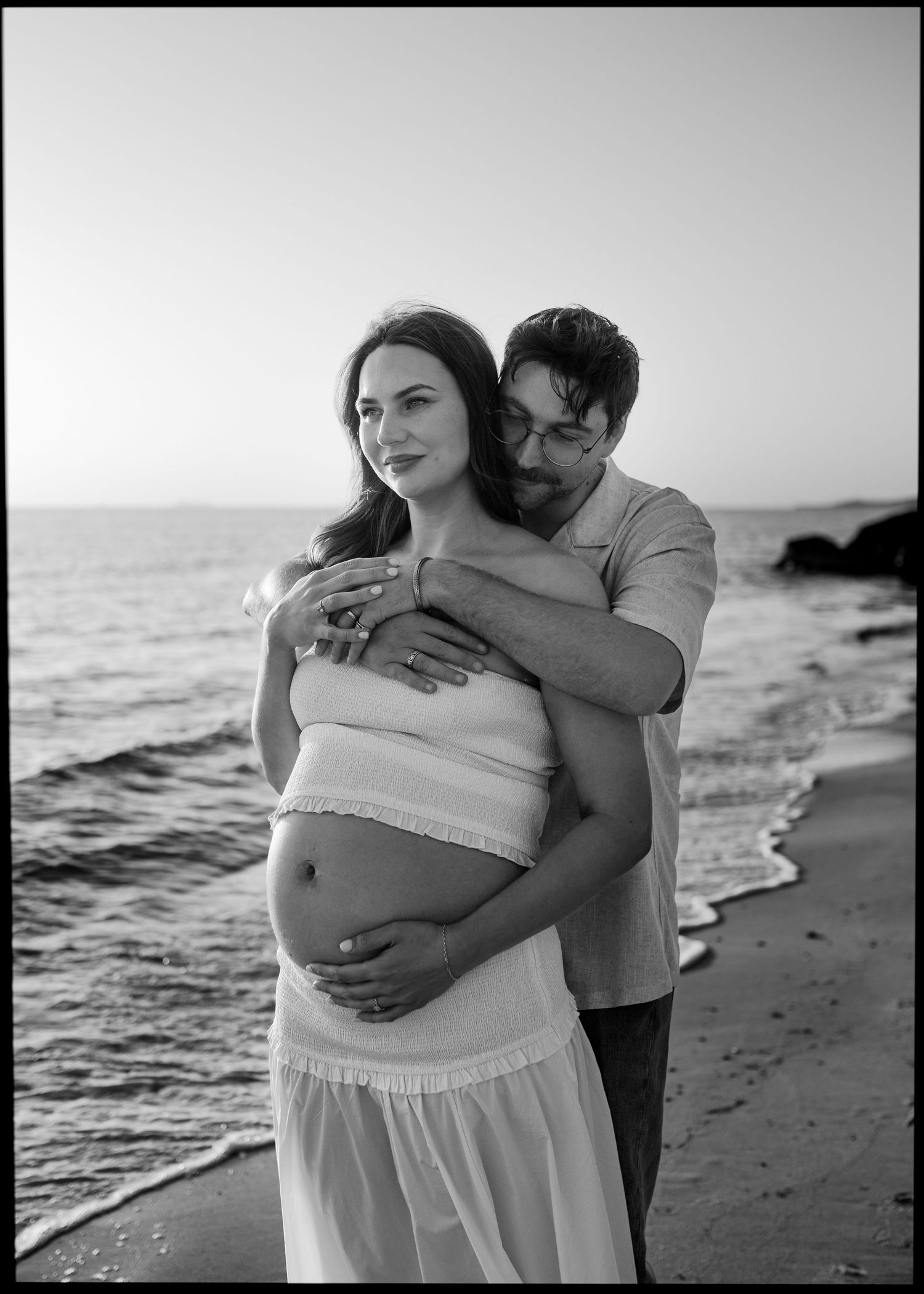 Black and white beach maternity photo of a couple, man embracing pregnant woman by the ocean shore.