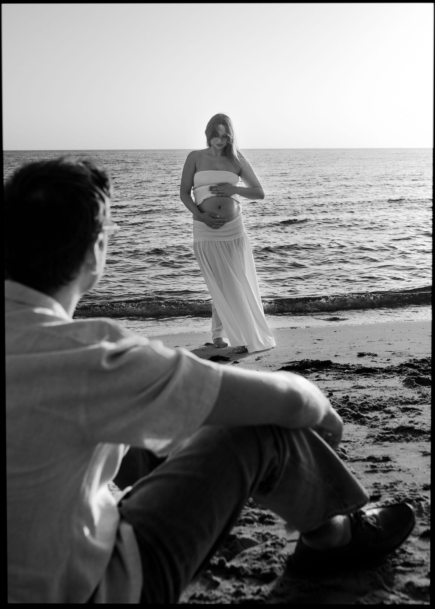 Black and white photo of pregnant woman in white dress on beach while man sits in foreground.