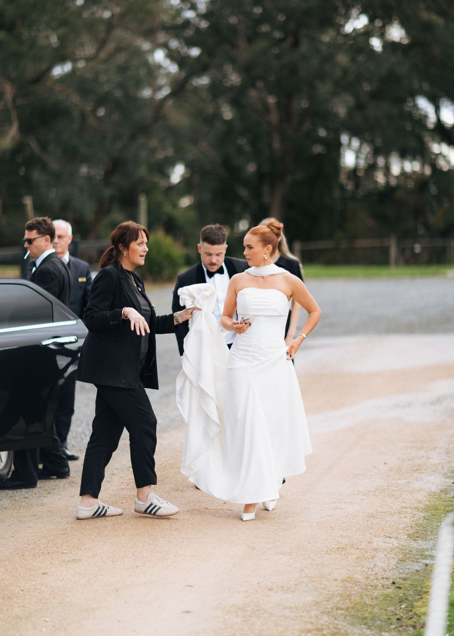 Wedding coordinator assists bride in white gown and groom on gravel driveway outside countryside venue.
