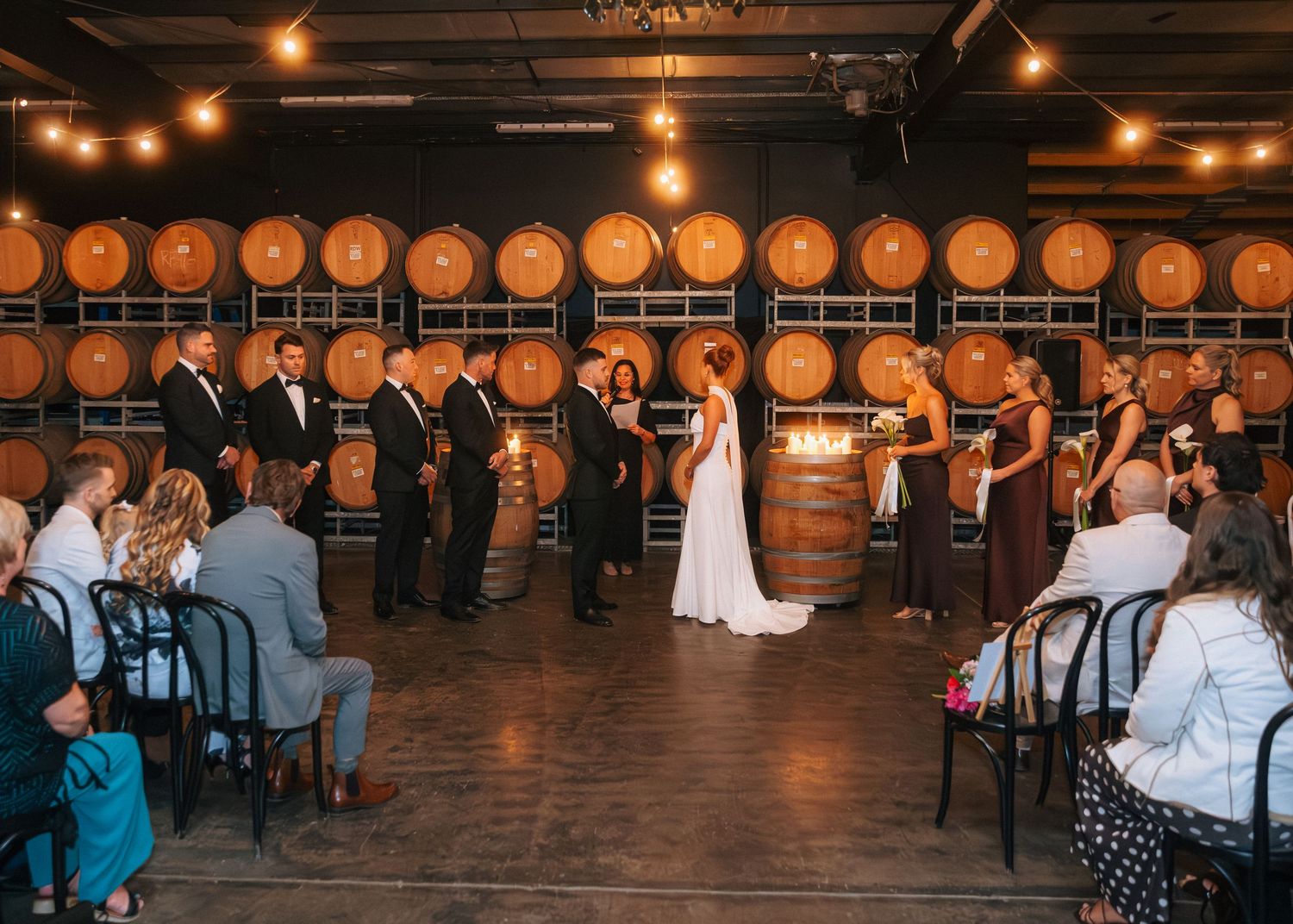 Wedding ceremony in a rustic winery with barrel wall backdrop, string lights, and guests seated in black chairs.