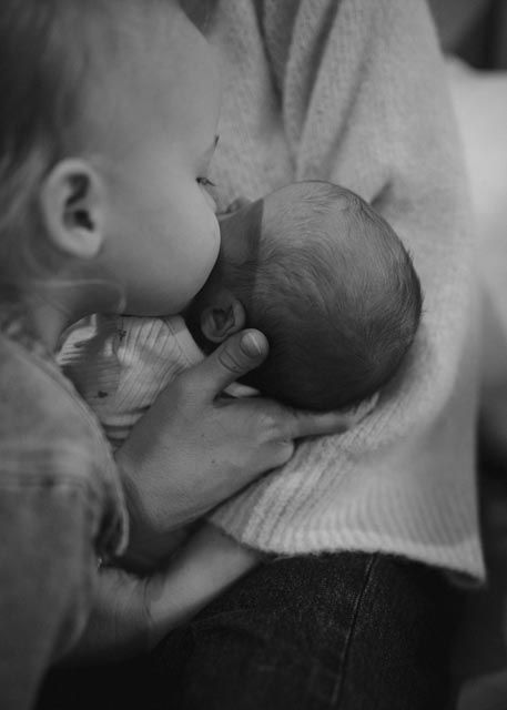Black and white photo of toddler sibling gently kissing newborn baby held by mother during family photoshoot.
