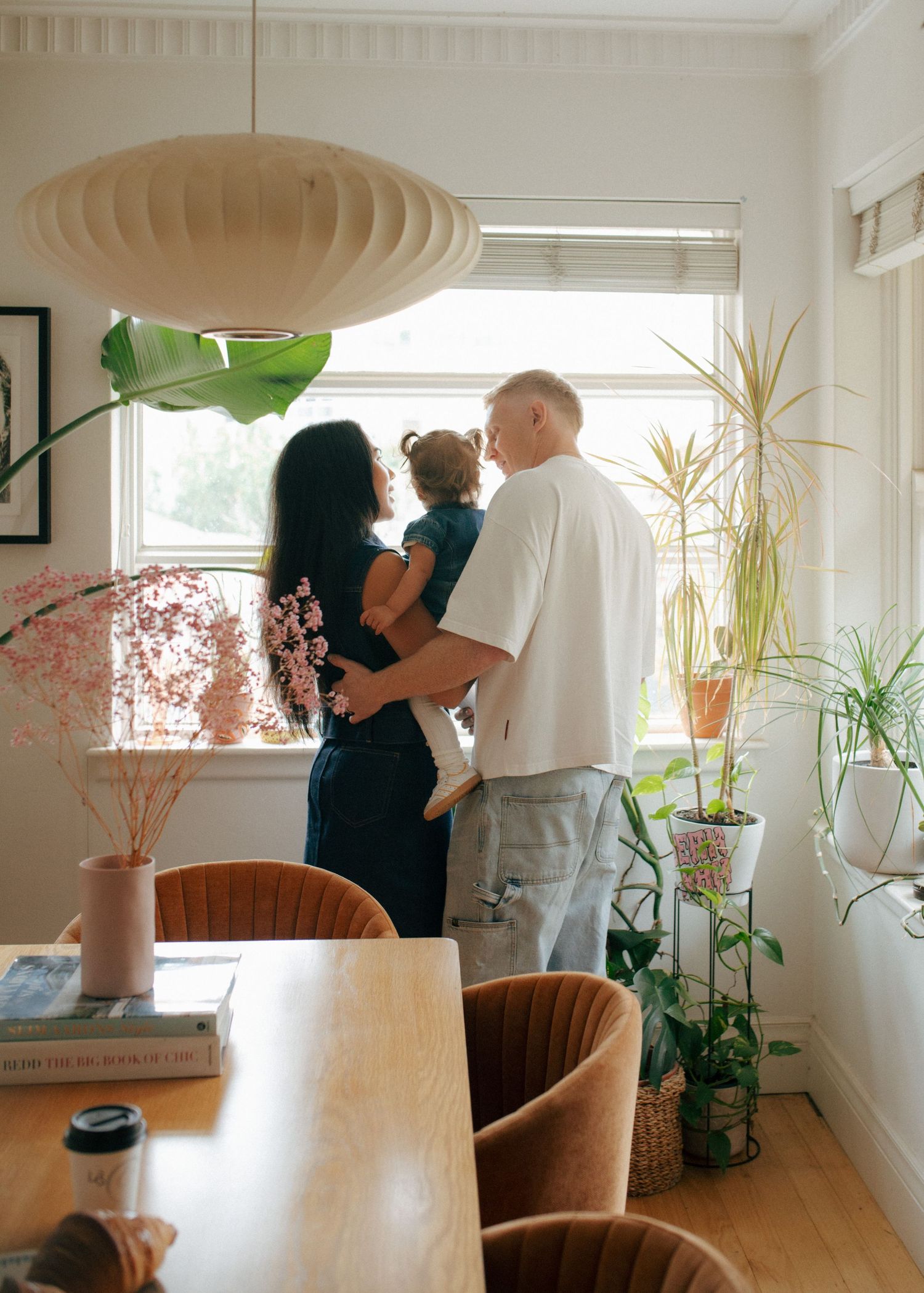 Parents holding baby while looking out window in plant-filled sunlit dining room.