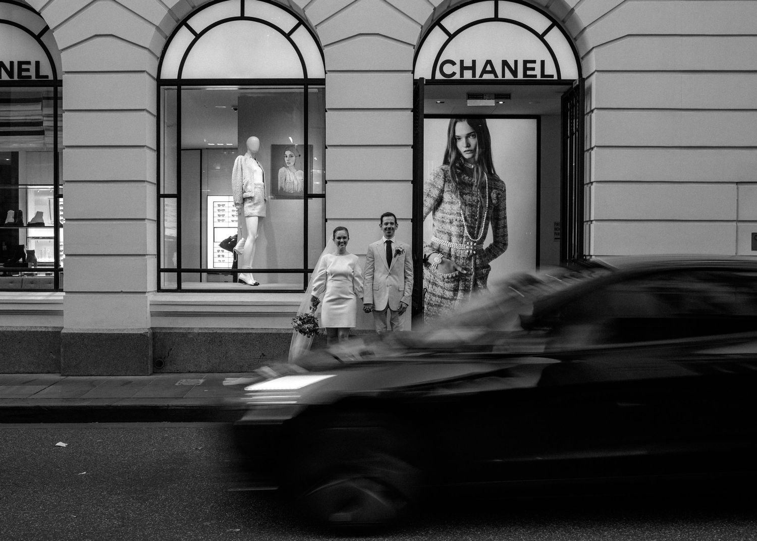 Black and white photo of wedding couple outside Chanel store with car passing on city street.
