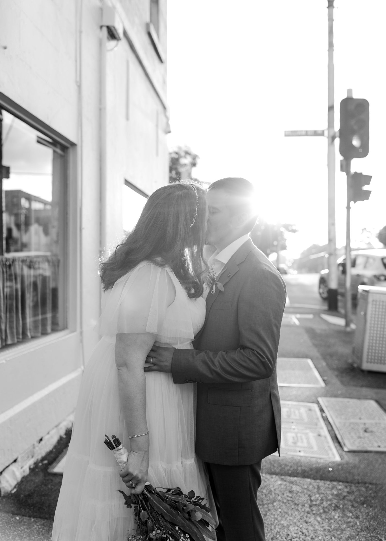 Black and white photo of bride and groom sharing a kiss on city street in golden backlight.