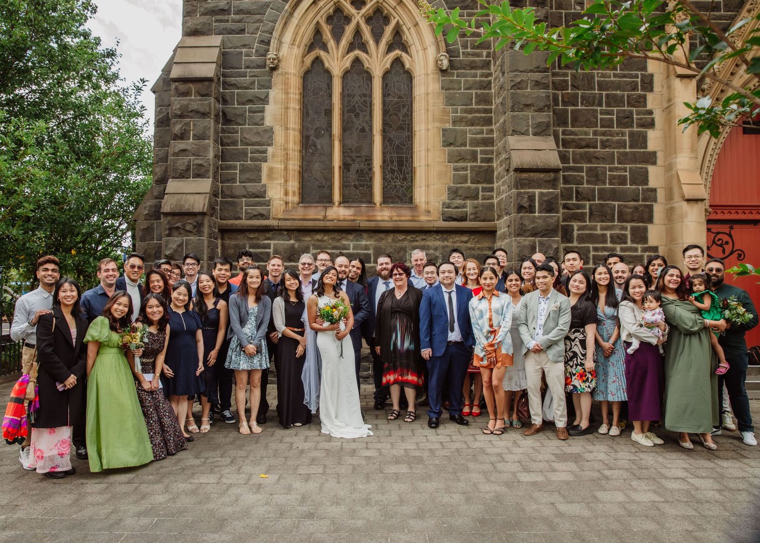 Large wedding group photo outside a Gothic stone church with bride and groom surrounded by guests.