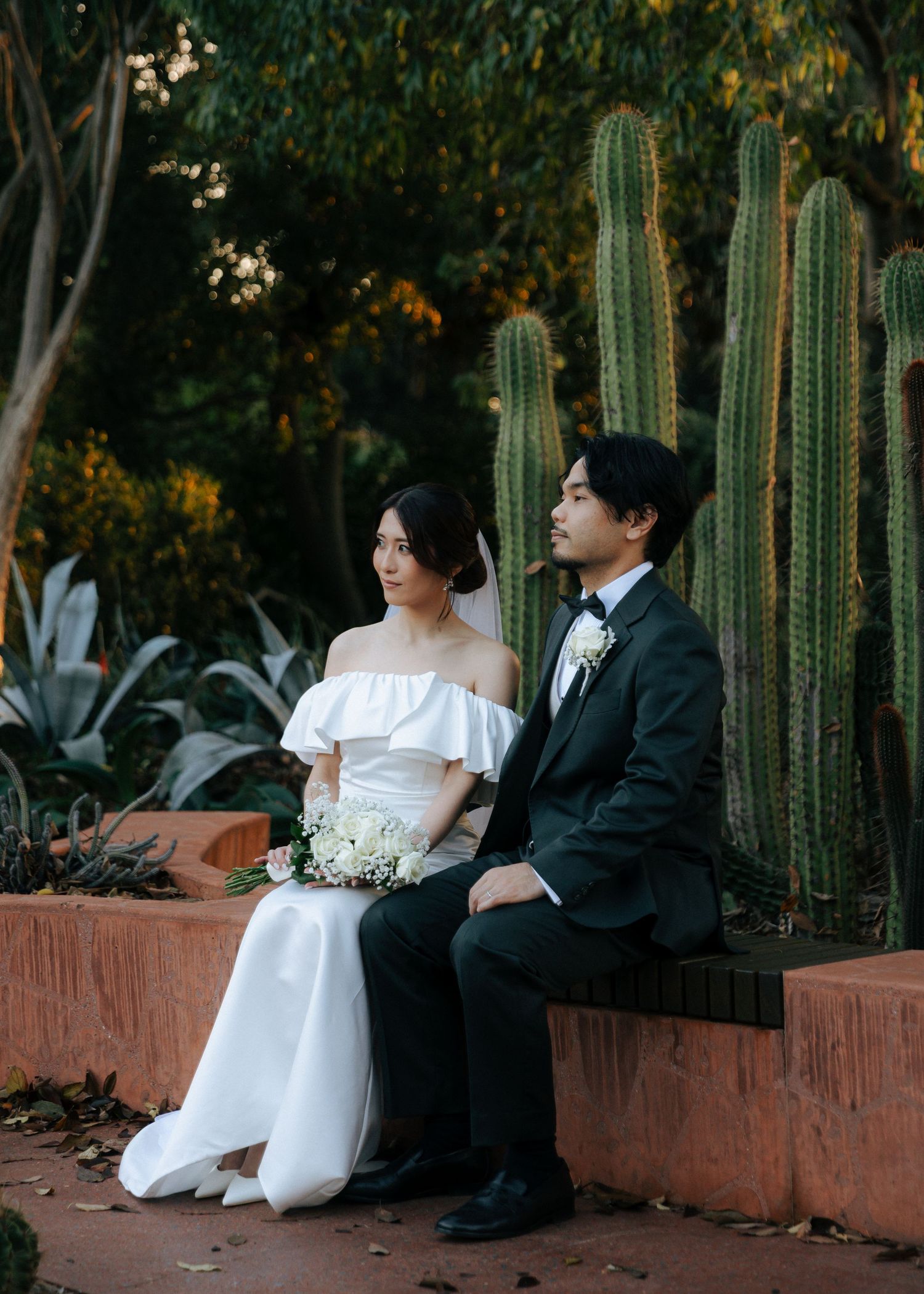 Bride and groom sitting together in a lush garden with tall cacti, bride holding white floral bouquet.