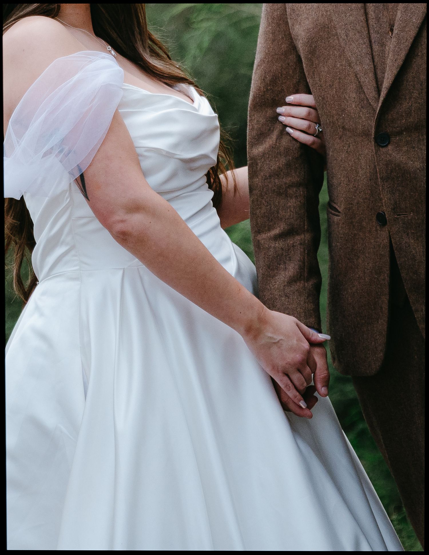 Bride in white off-shoulder gown holds hands with groom in brown tweed suit during outdoor wedding ceremony.