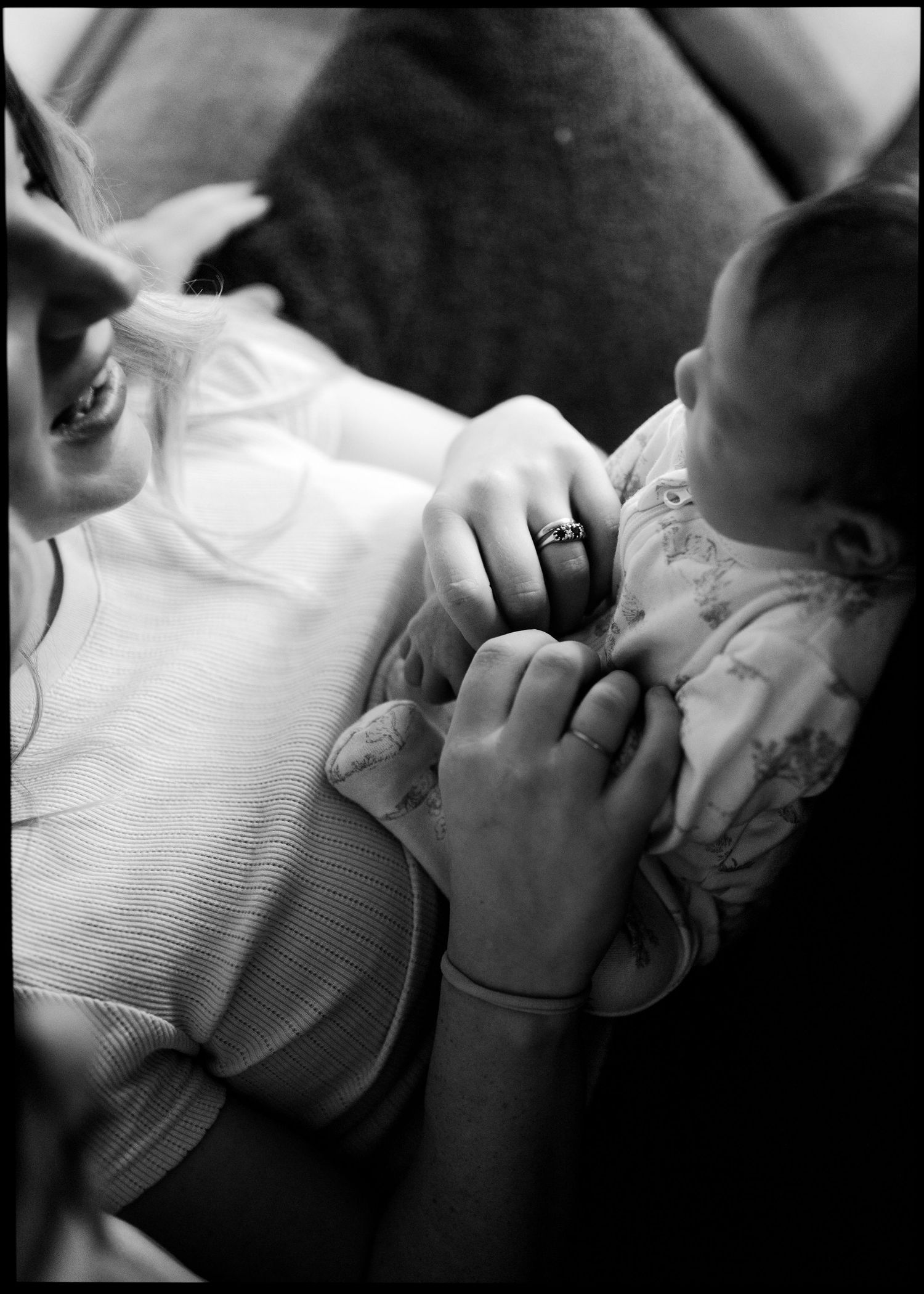 Black and white photo of a smiling mother in a ribbed top cradling her newborn baby in a floral outfit, with a wedding ring on her hand.