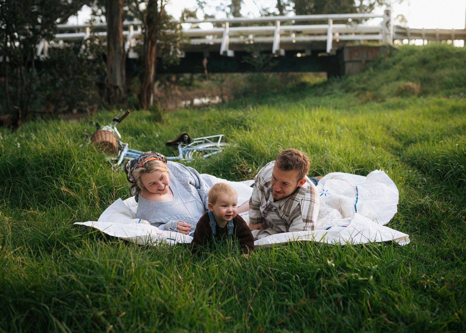 Happy young family with toddler relaxing on a white blanket in lush green grass outdoors.