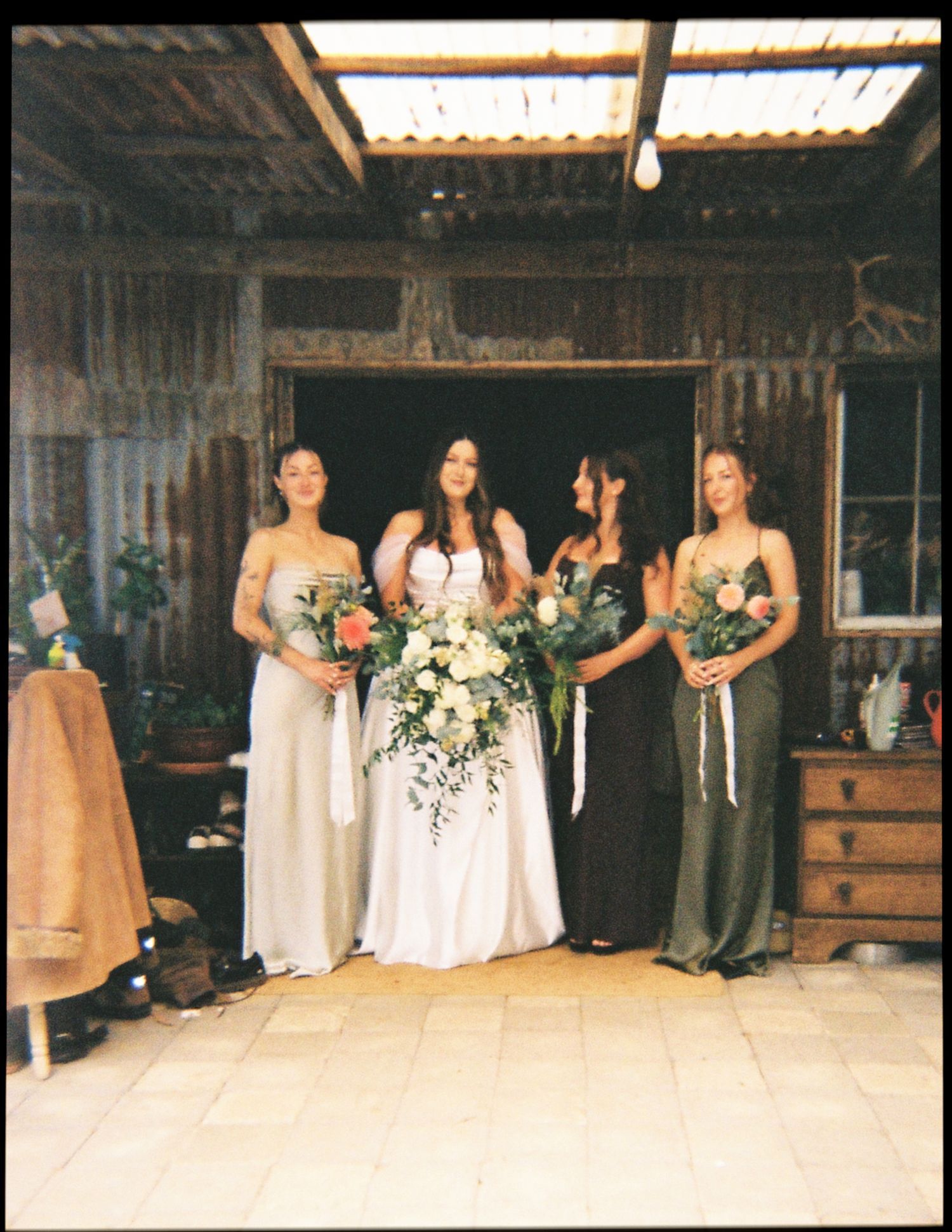 Bride in white gown with three bridesmaids in muted tones holding bouquets inside a rustic barn venue.