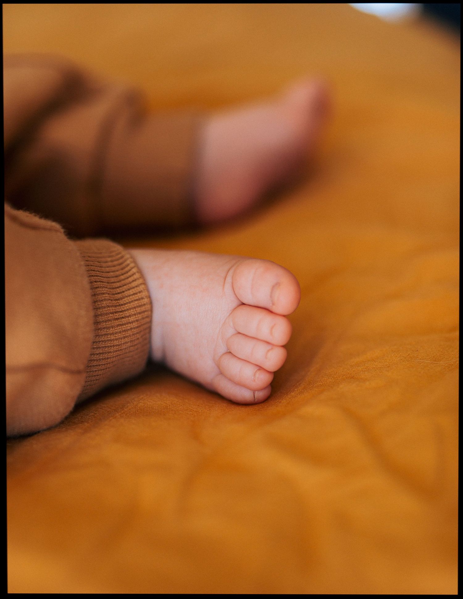 Close-up of a baby's tiny bare foot in a brown outfit resting on a warm mustard-yellow blanket, with the other foot softly blurred behind.