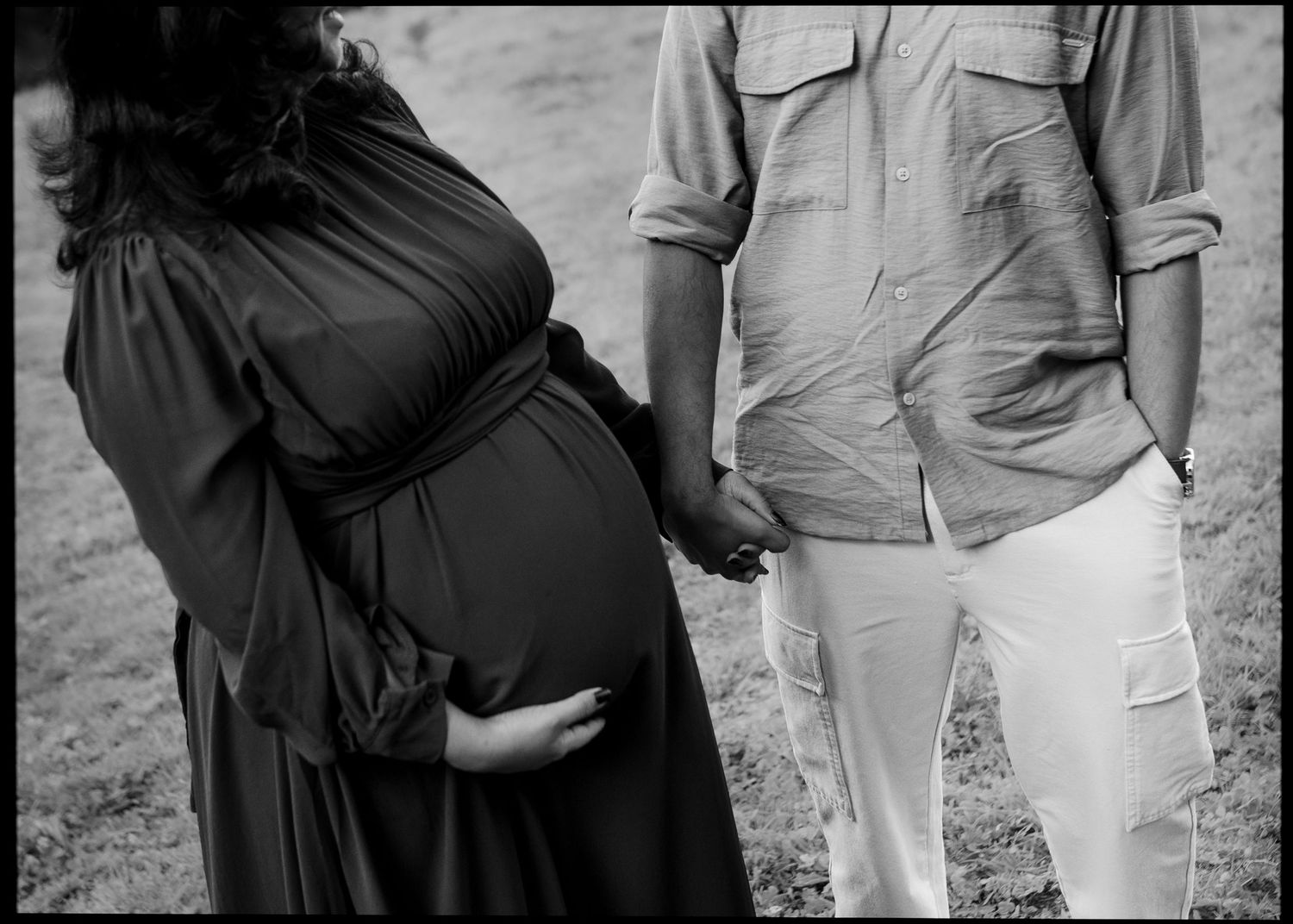 Black and white maternity photo of a pregnant woman in a flowing dress holding hands with her partner in a button-up shirt and cargo pants.