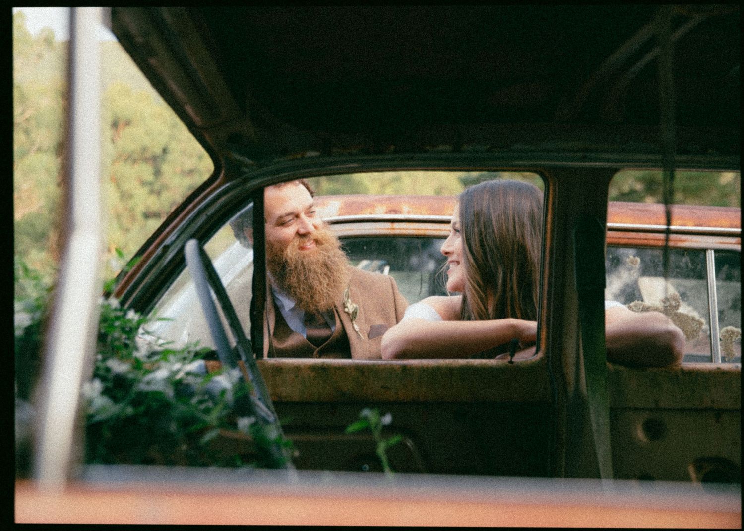 Bearded groom and bride share an intimate moment viewed through the window of a rusty vintage car.