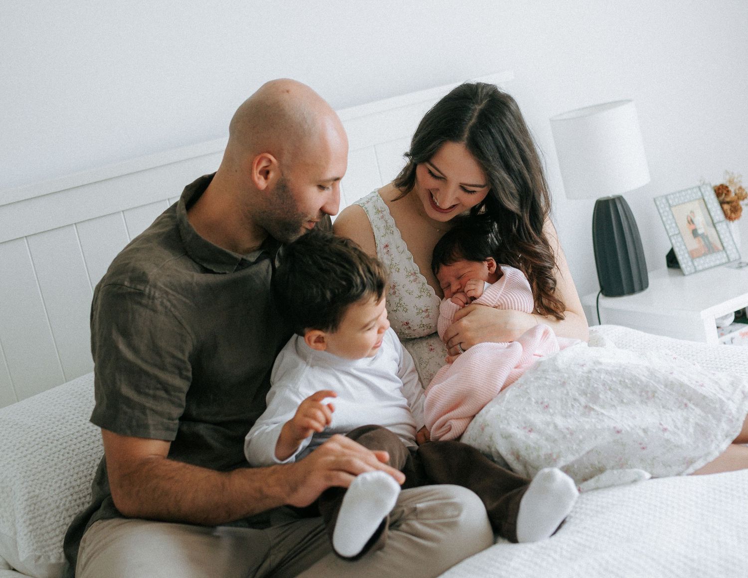 Family of four on bed, parents gazing lovingly at toddler and newborn baby girl in pink.