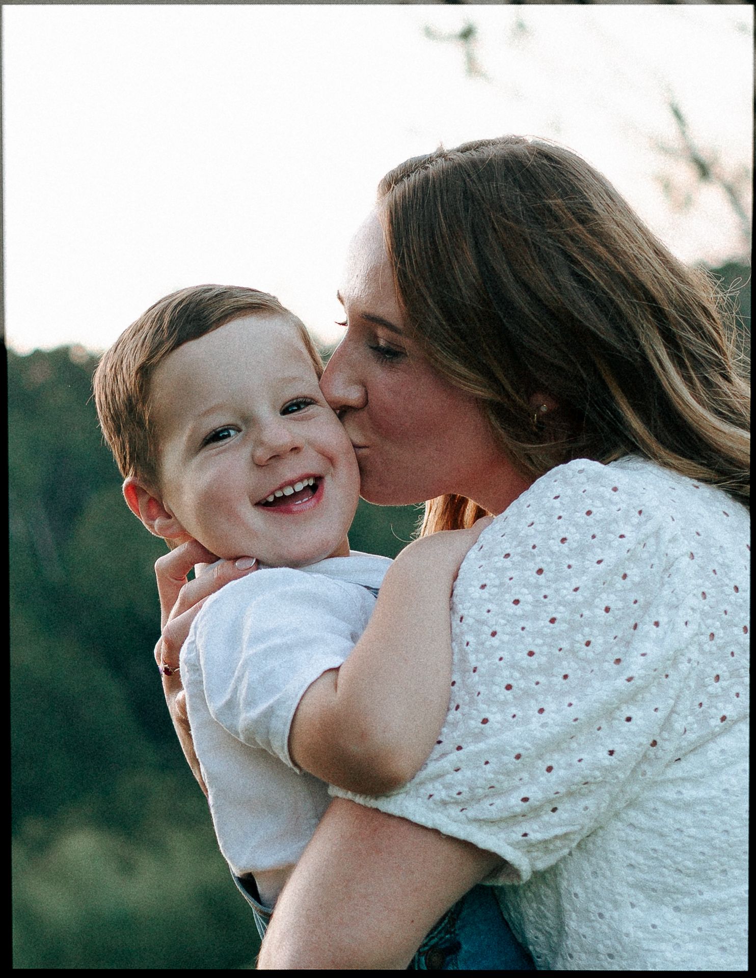 A woman kisses a laughing toddler boy on the cheek in a warm outdoor portrait.