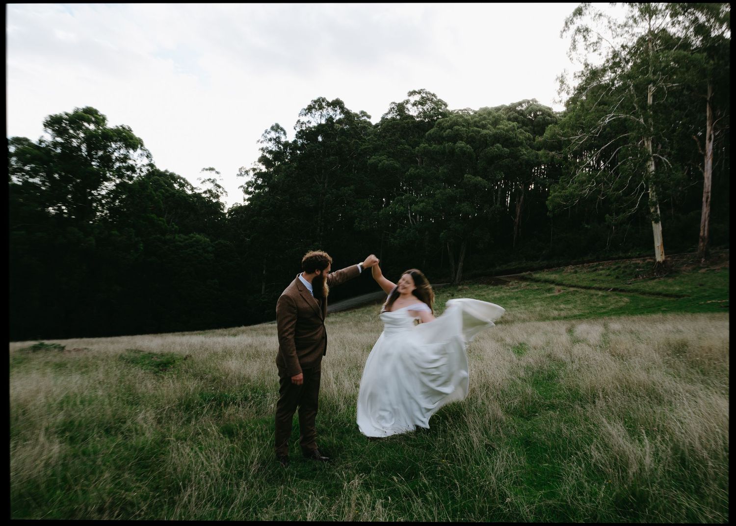 Groom twirls bride in white gown in open grassy forest clearing.