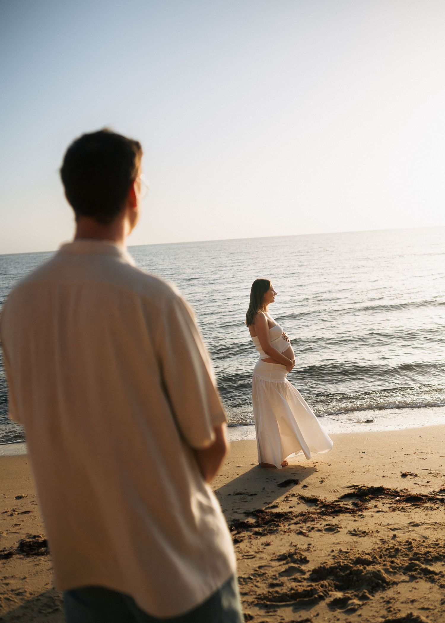 Pregnant woman in white dress stands at ocean shore while man watches from behind on sandy beach.