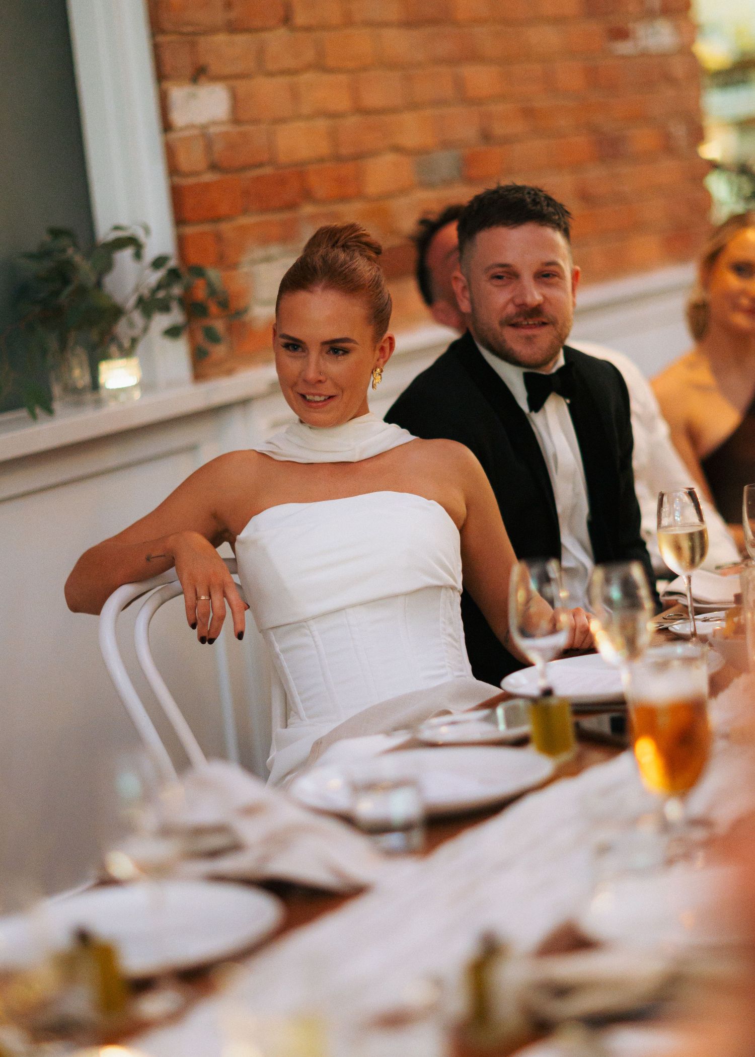 Bride in white strapless gown and groom in tuxedo seated at wedding reception table with guests.