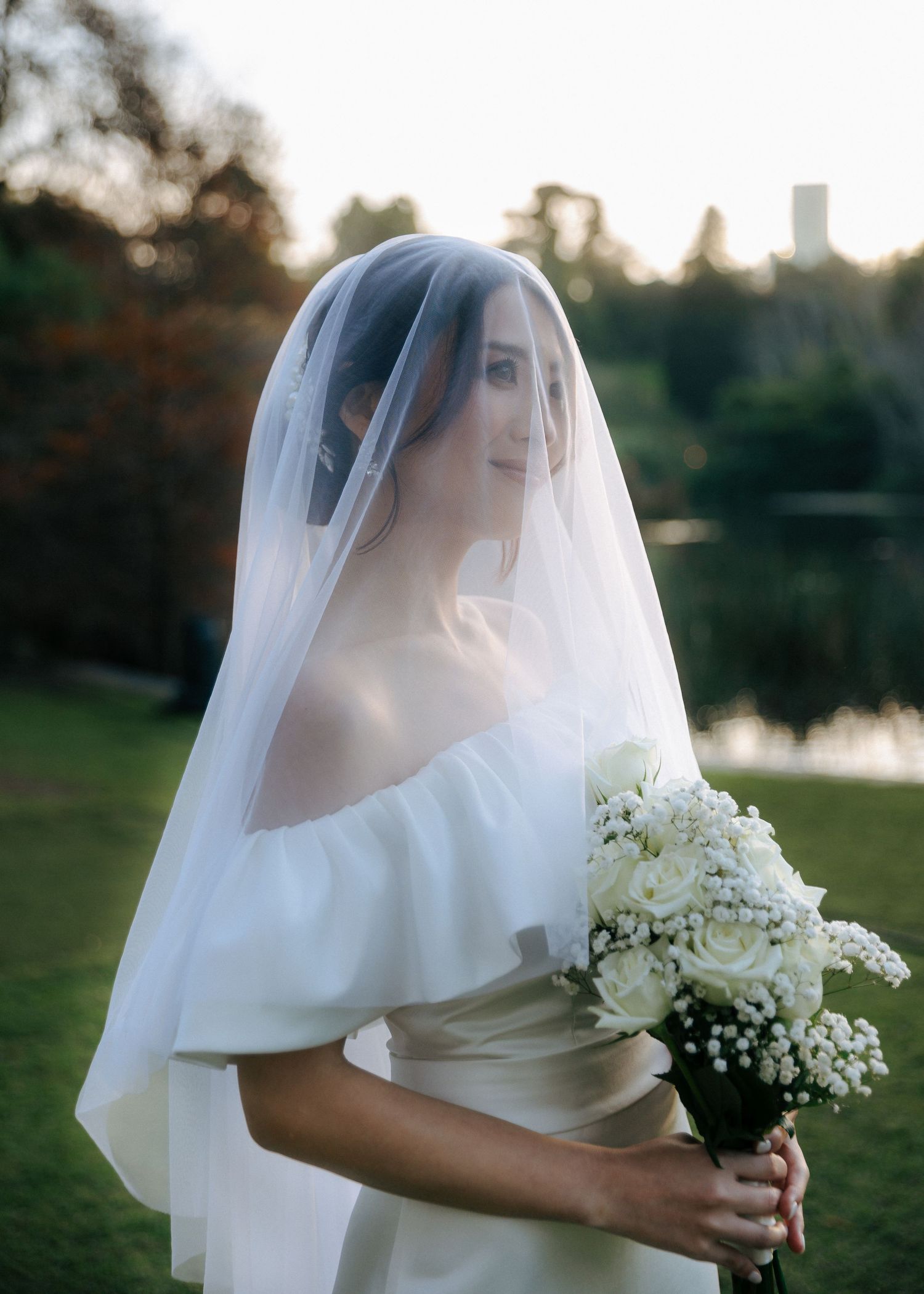 Elegant bride wearing a flowing veil and off-shoulder gown holds a white bouquet at golden hour outdoors.