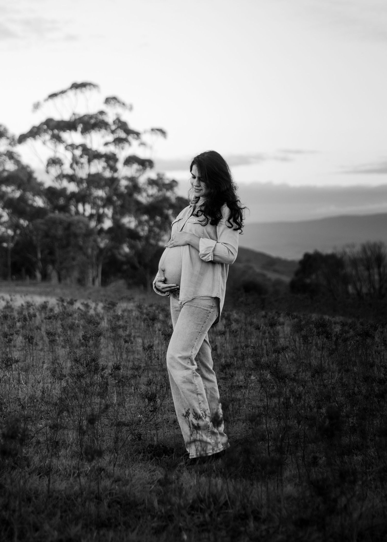 Black and white portrait of a pregnant woman standing alone in a field at dusk, cradling her baby bump.