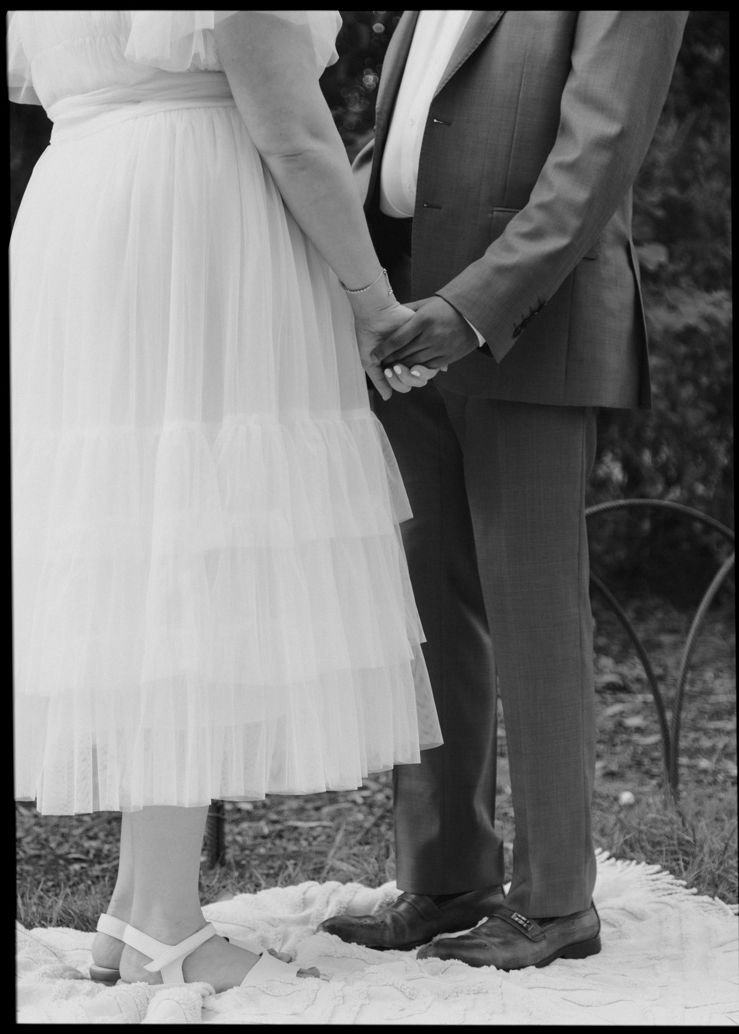 Black and white close-up of bride and groom holding hands at wedding ceremony on white blanket.