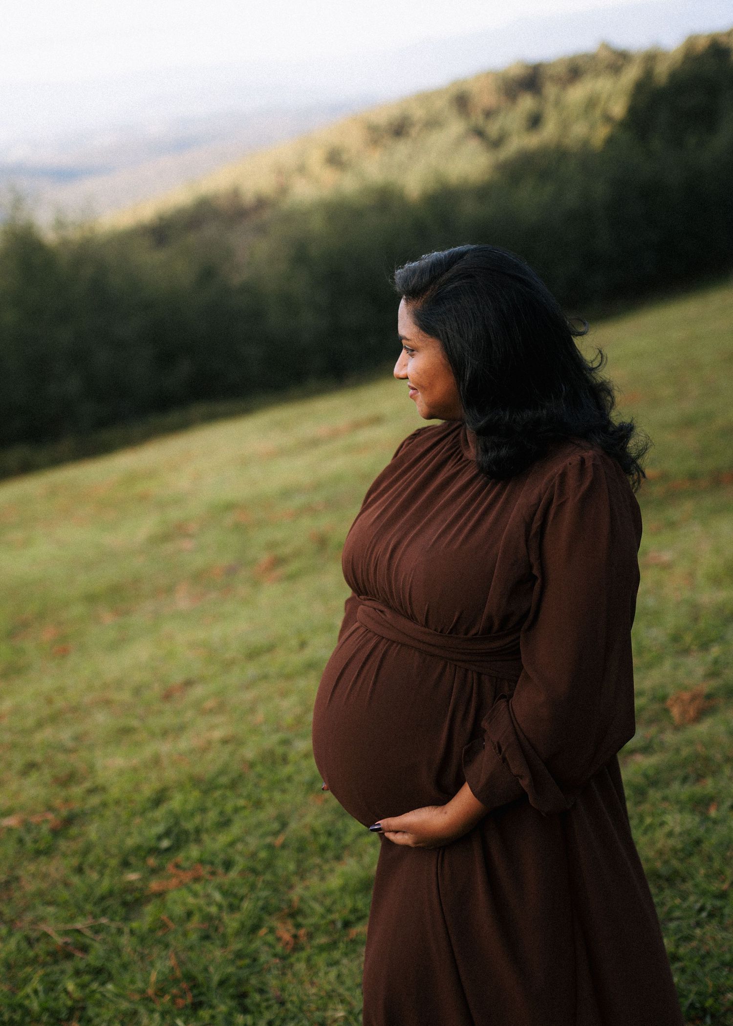 Pregnant woman in a flowing brown dress cradling her baby bump while standing on a grassy hillside at sunset.