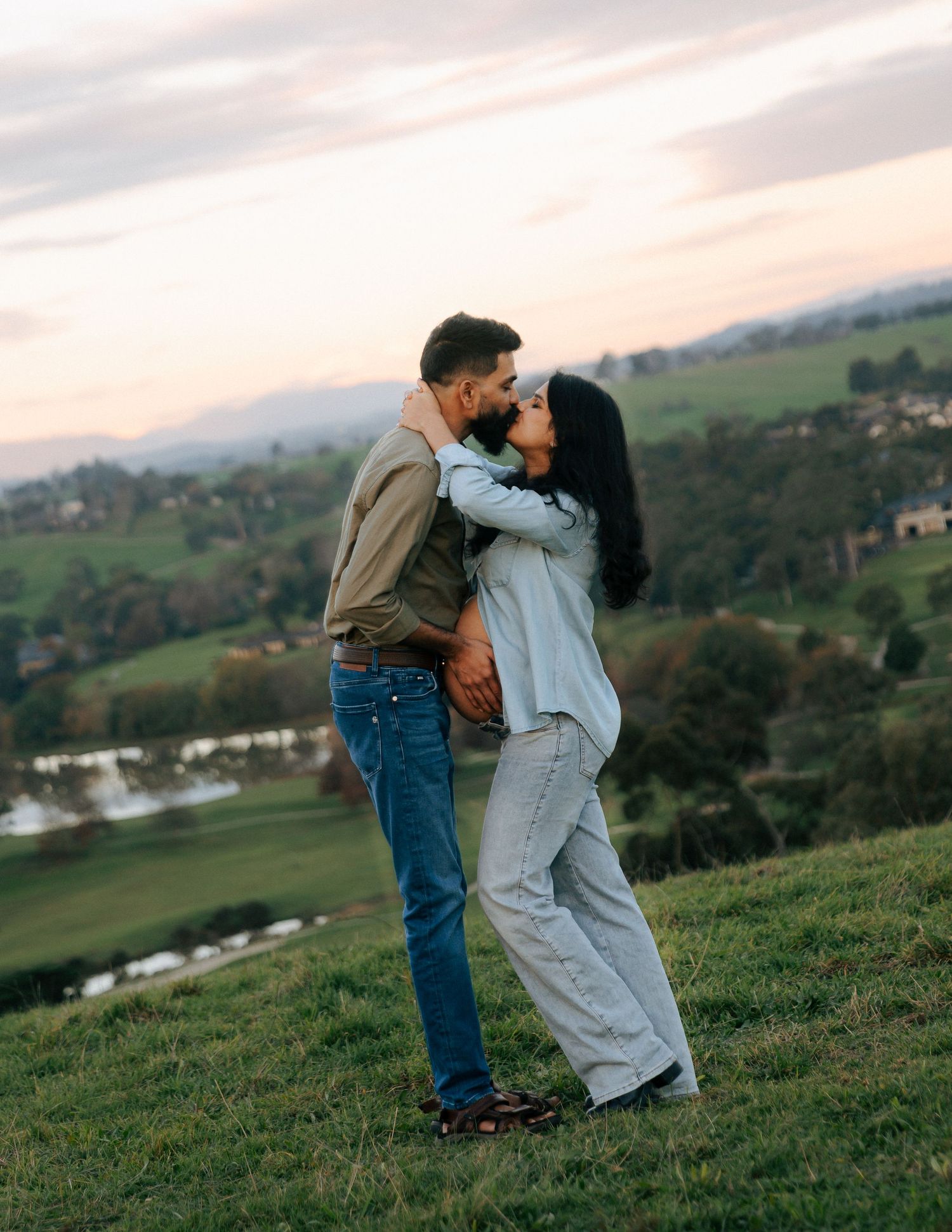 Couple sharing a romantic kiss on a lush green hillside during a beautiful sunset maternity photoshoot.