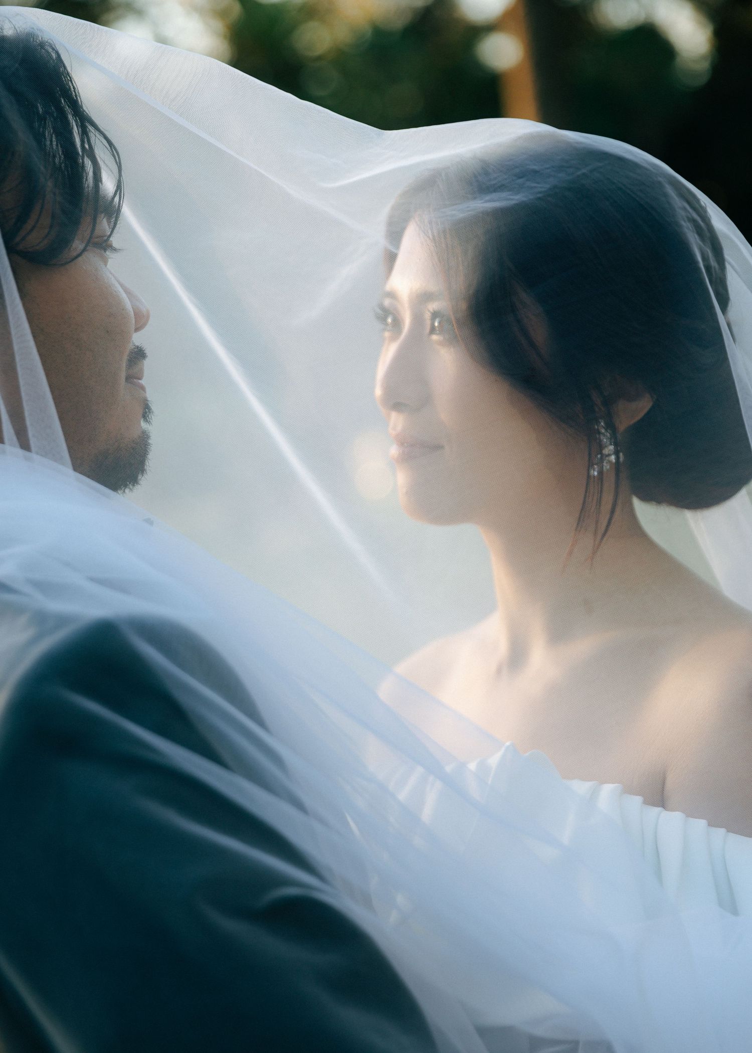 Romantic bride and groom portrait beneath a flowing white veil, softly lit in warm outdoor light.