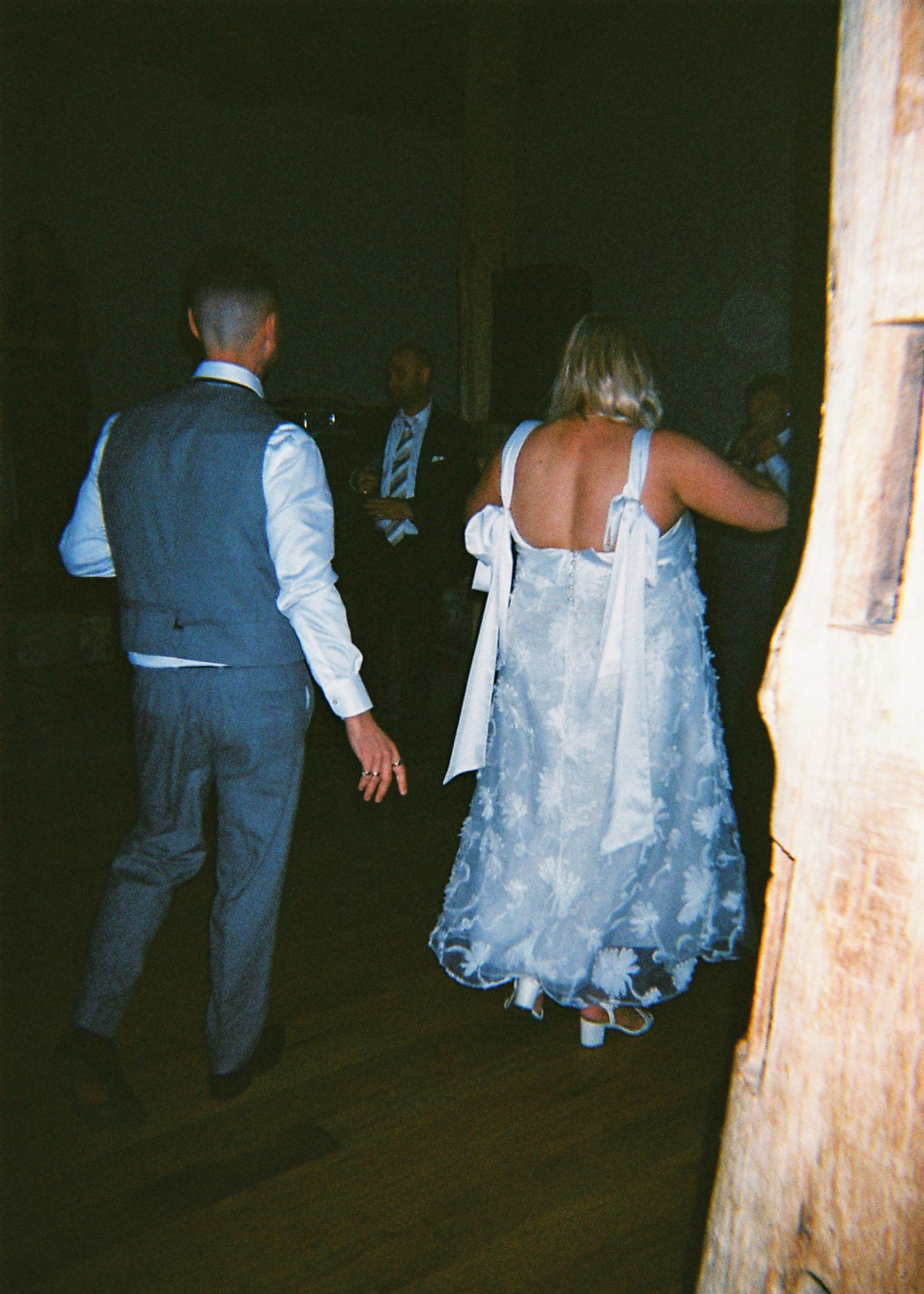 Bride in elegant backless lace gown with bow and groom walking hand in hand at their wedding reception.