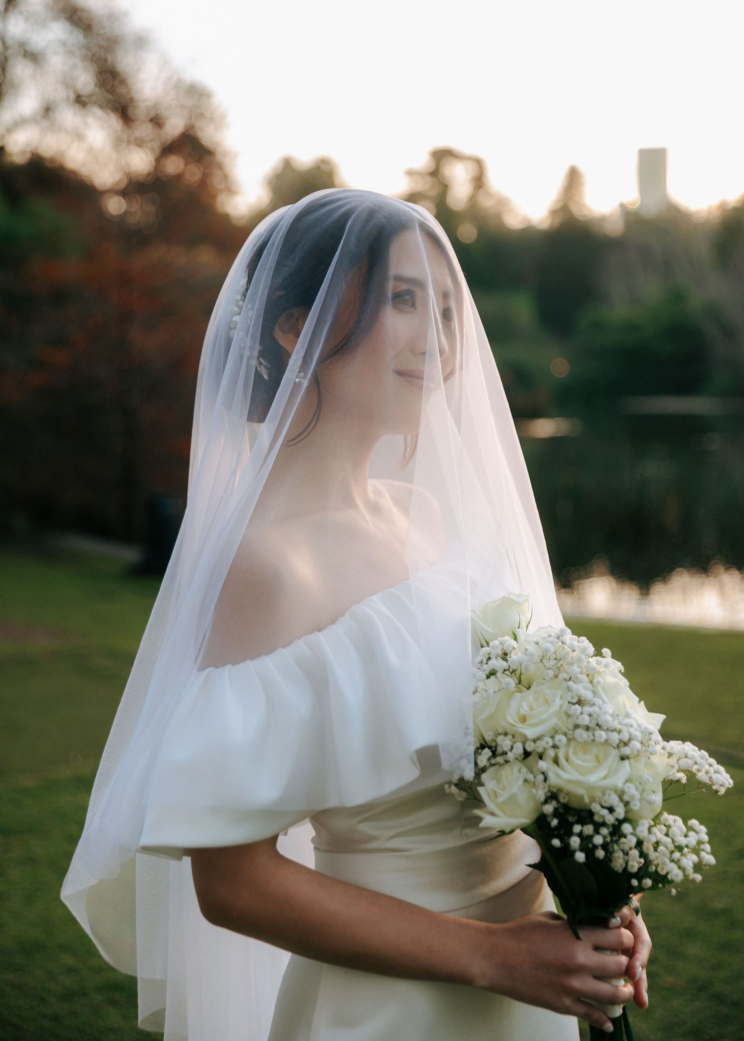 Bride in white off-shoulder gown and veil holds white rose bouquet outdoors near a lake at golden hour.
