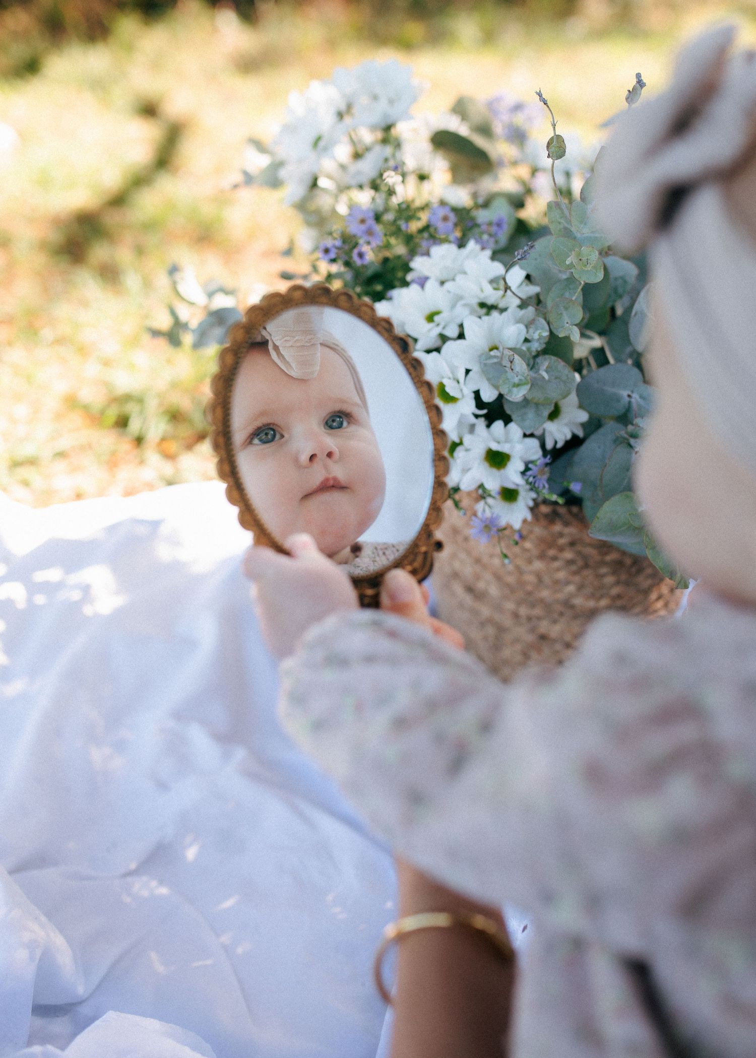 Baby girl with bow headband reflected in vintage hand mirror among flowers.
