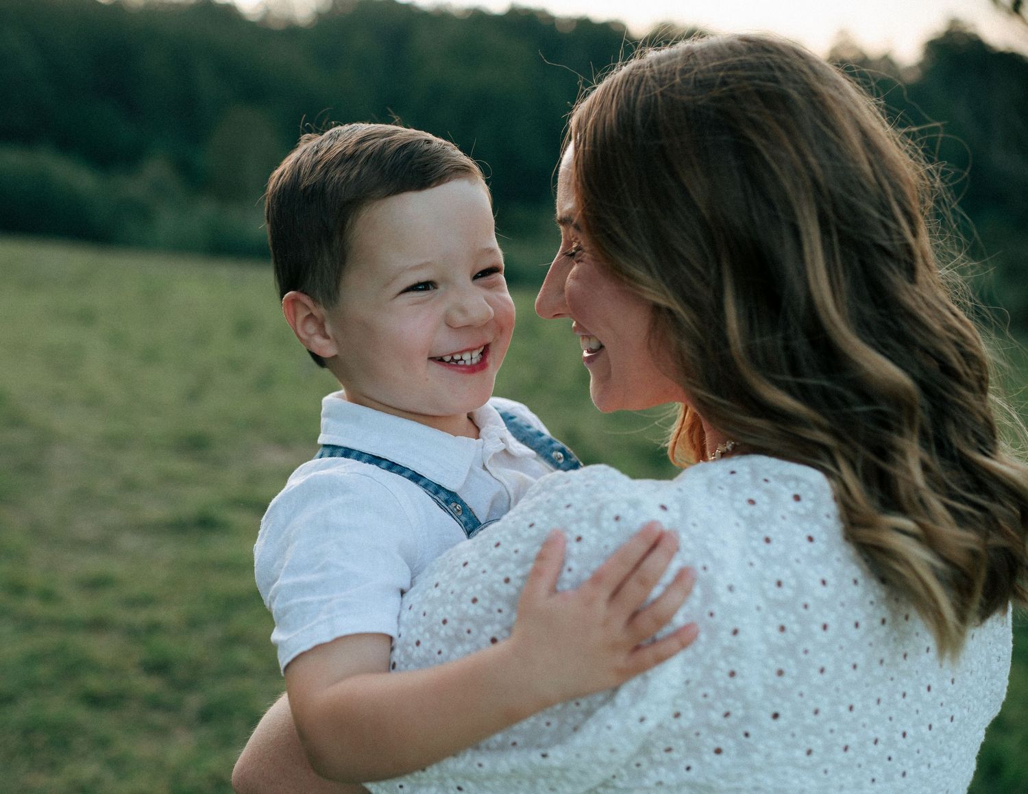 Smiling toddler boy in white shirt being held by his laughing mother in sunny green field.