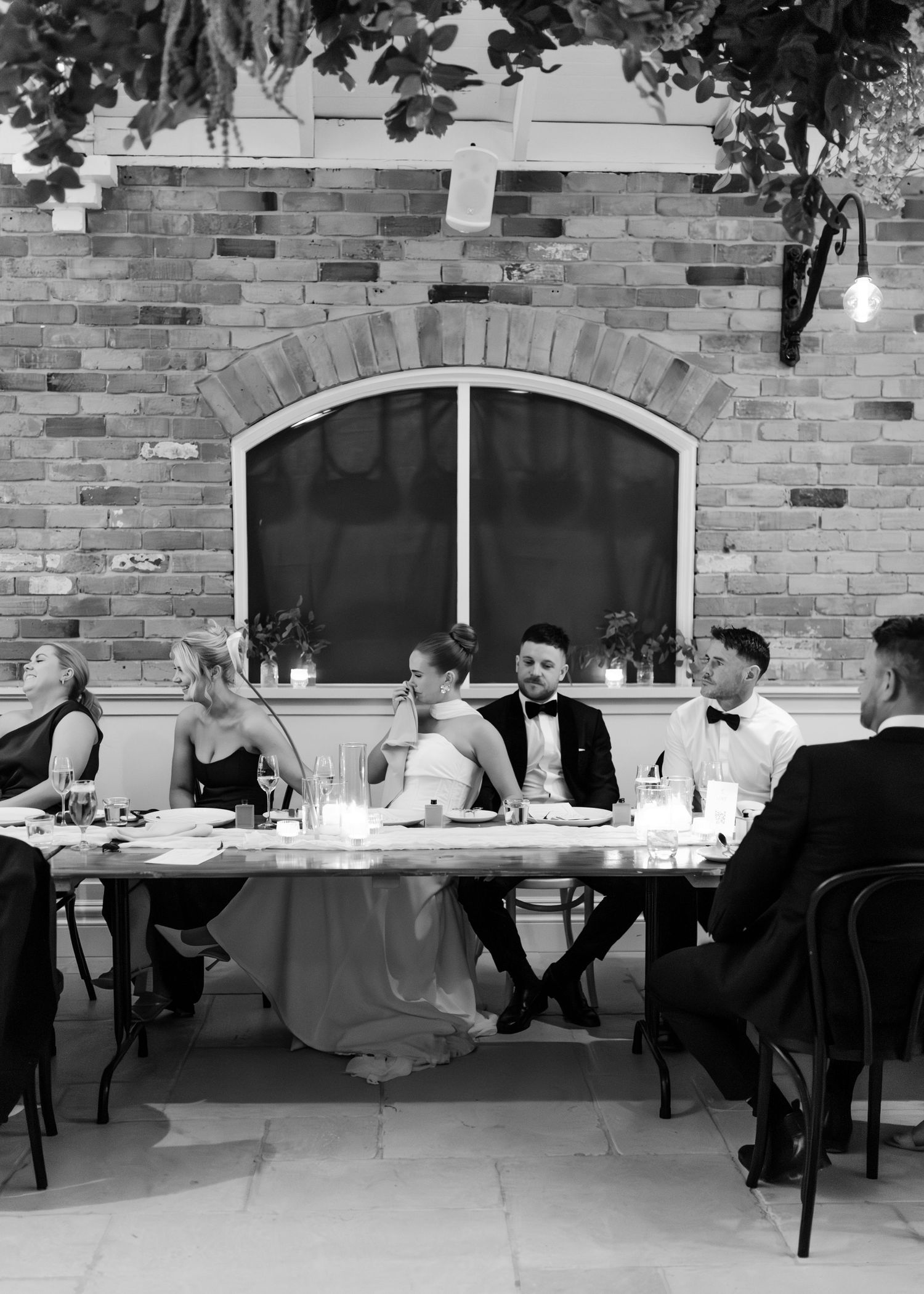 Black and white photo of wedding party seated at head table against brick wall with arched window.