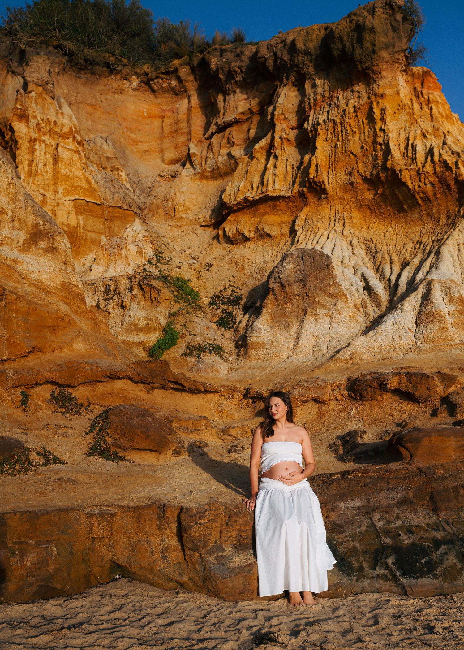 Pregnant woman in white bandeau top and skirt poses before dramatic golden sandstone cliffs at beach.