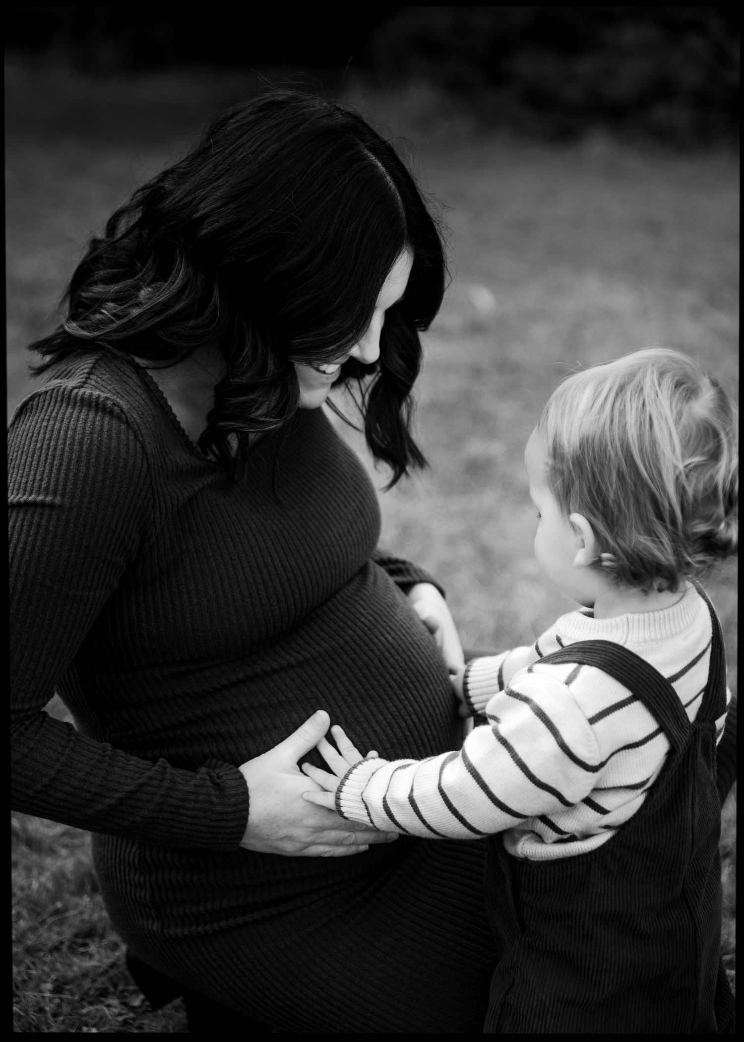 Black and white photo of pregnant mother and toddler sharing tender moment outdoors.